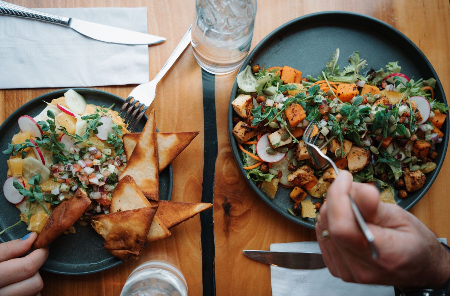 Two dark plates of fresh salads with greens and vegetables shared by diners at a wooden restaurant table.