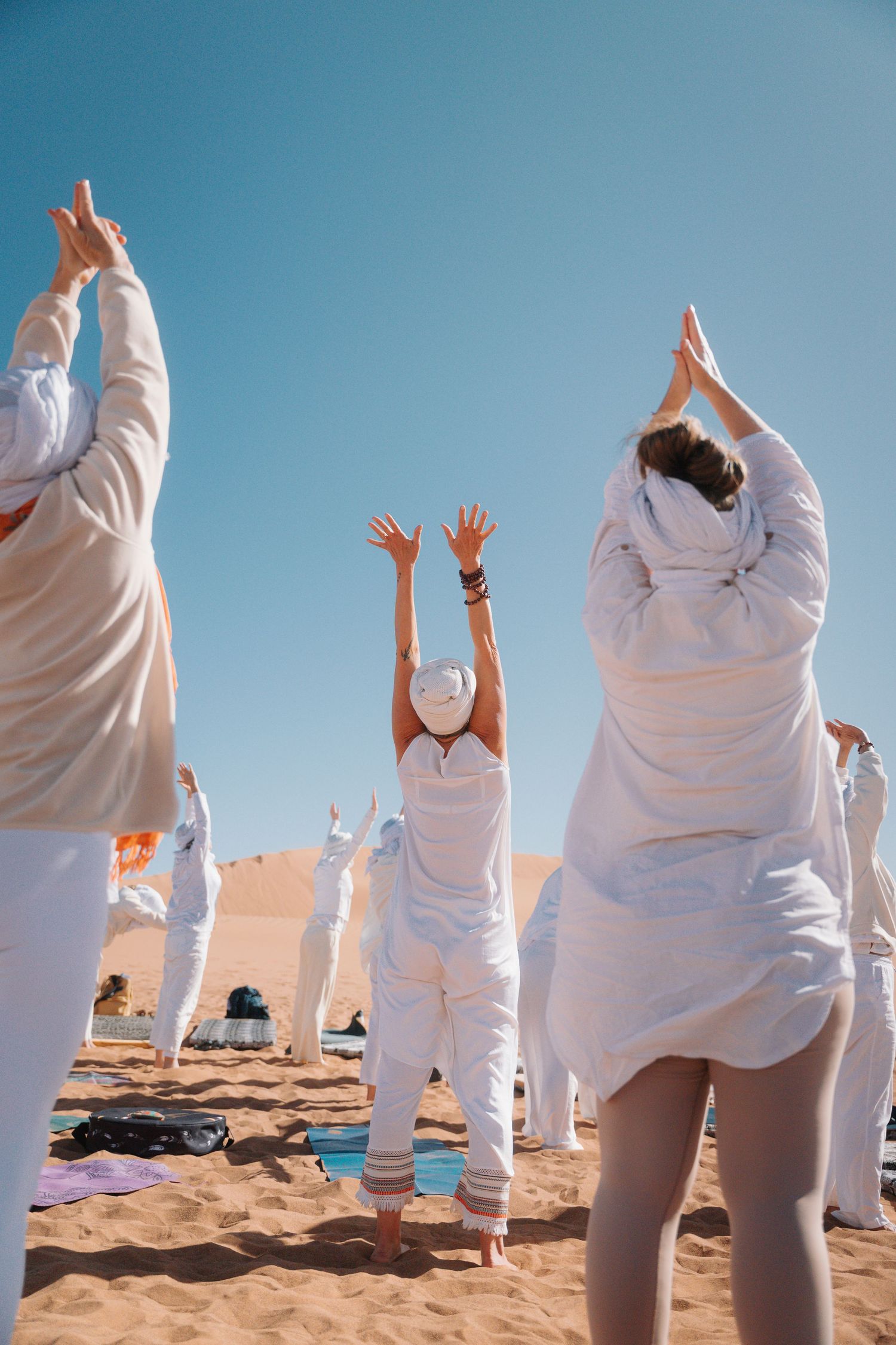 Group of women in white clothing practicing yoga with arms raised on desert sand dunes.