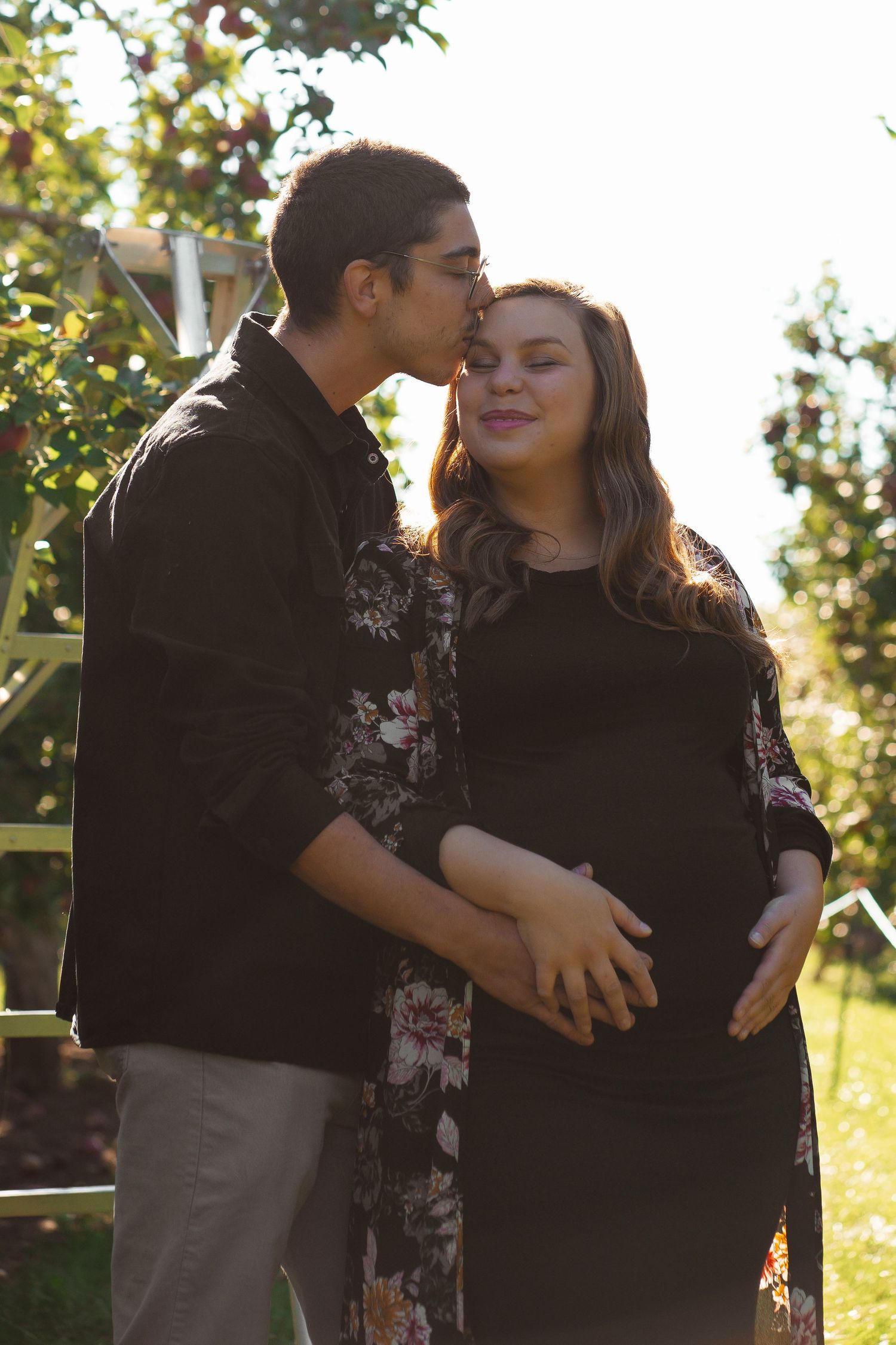 A man kissing a pregnant woman on the forehead in a sunlit orchard.
