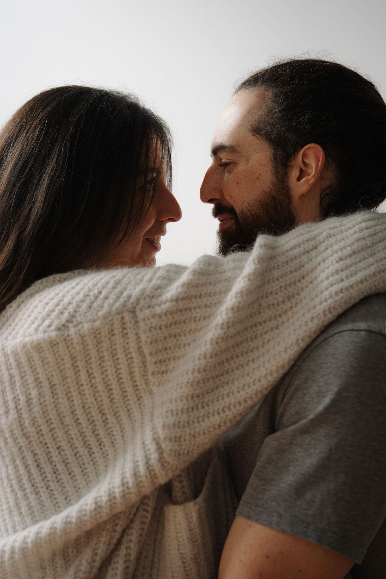 Romantic couple wrapped in cozy white knit blanket, smiling face to face against white background.