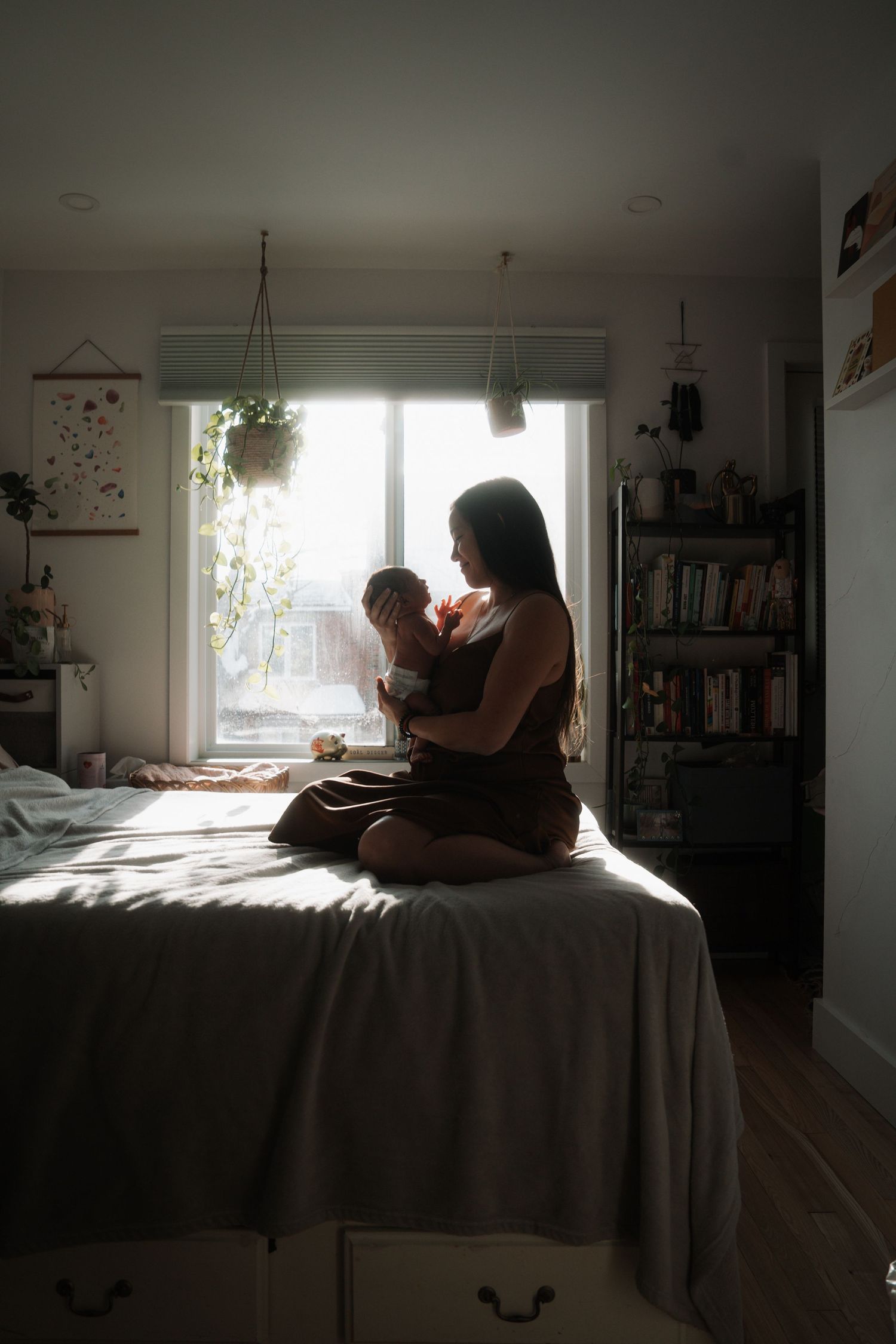 Mother sitting cross-legged on bed holding newborn in softly lit bedroom.