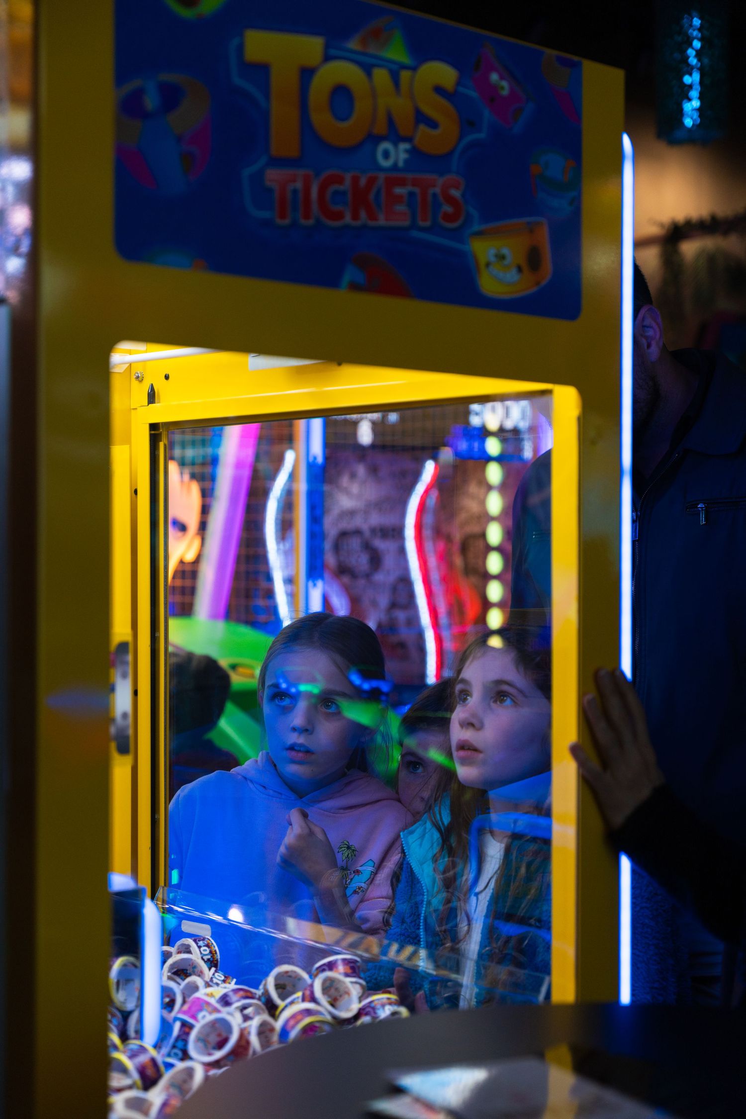Two children peer through the glass of a yellow Tons of Tickets arcade prize machine.