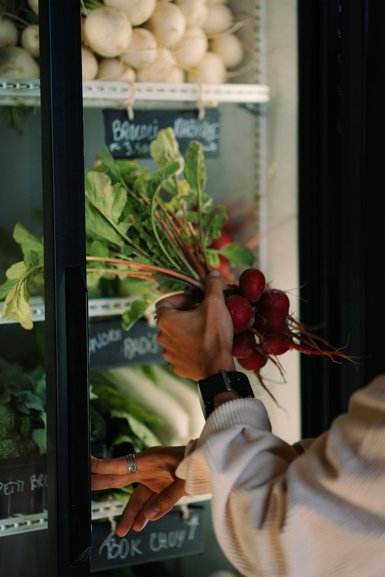 Person holding a fresh bunch of red beets near an open refrigerator display at a specialty grocery store.