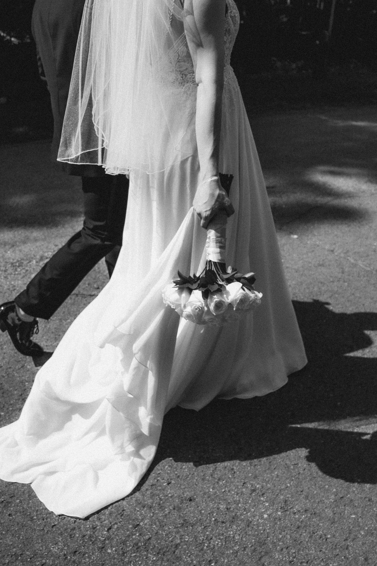 Black and white photo of a bride in a flowing white gown holding a bouquet while walking with a groom.
