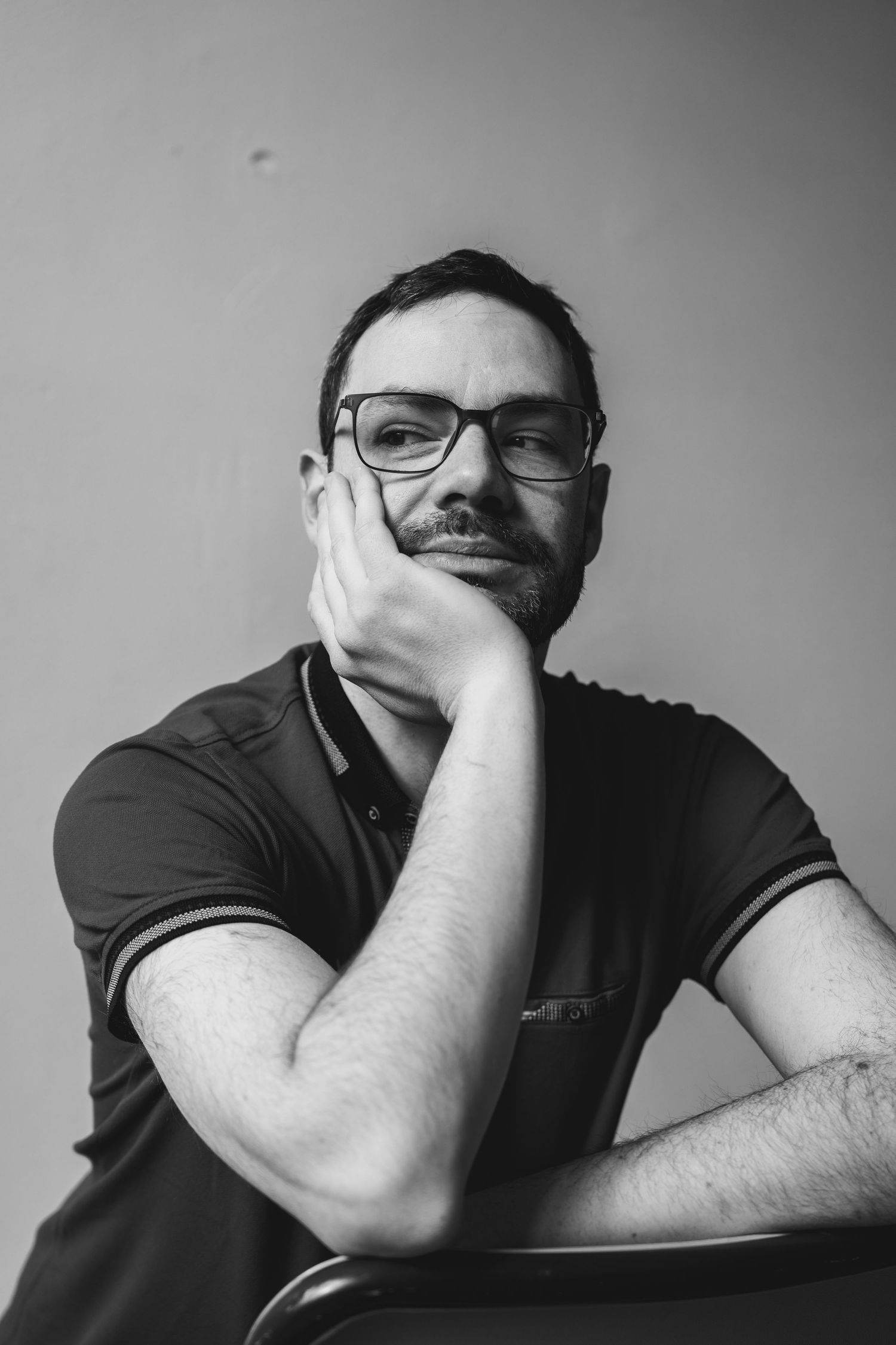 Thoughtful bearded man with glasses resting chin on hand in black polo.