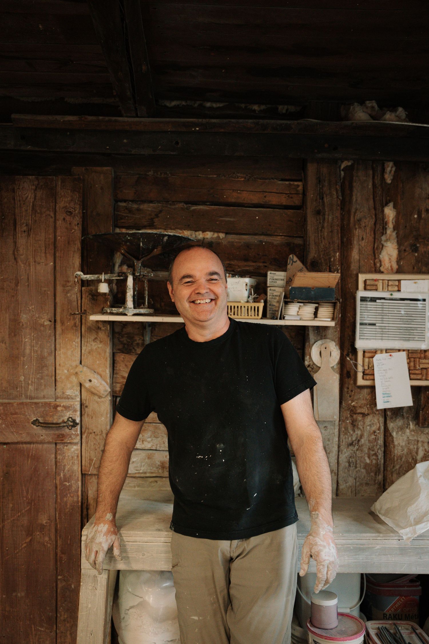 Smiling craftsman in black t-shirt with clay-covered hands stands in rustic wooden workshop.