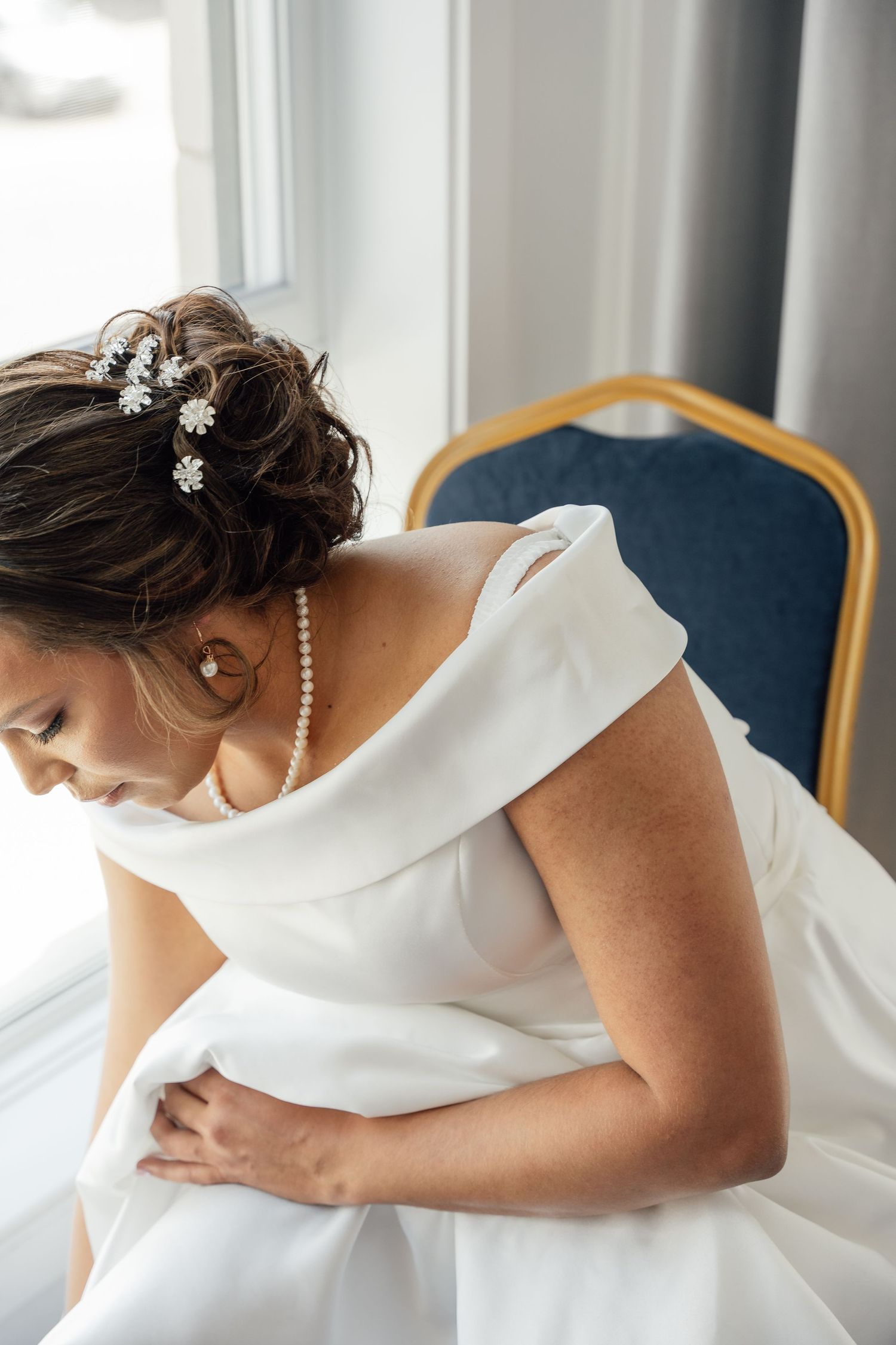 An elegant bride in an off-shoulder white gown with floral hair accessories sits pensively by a window.