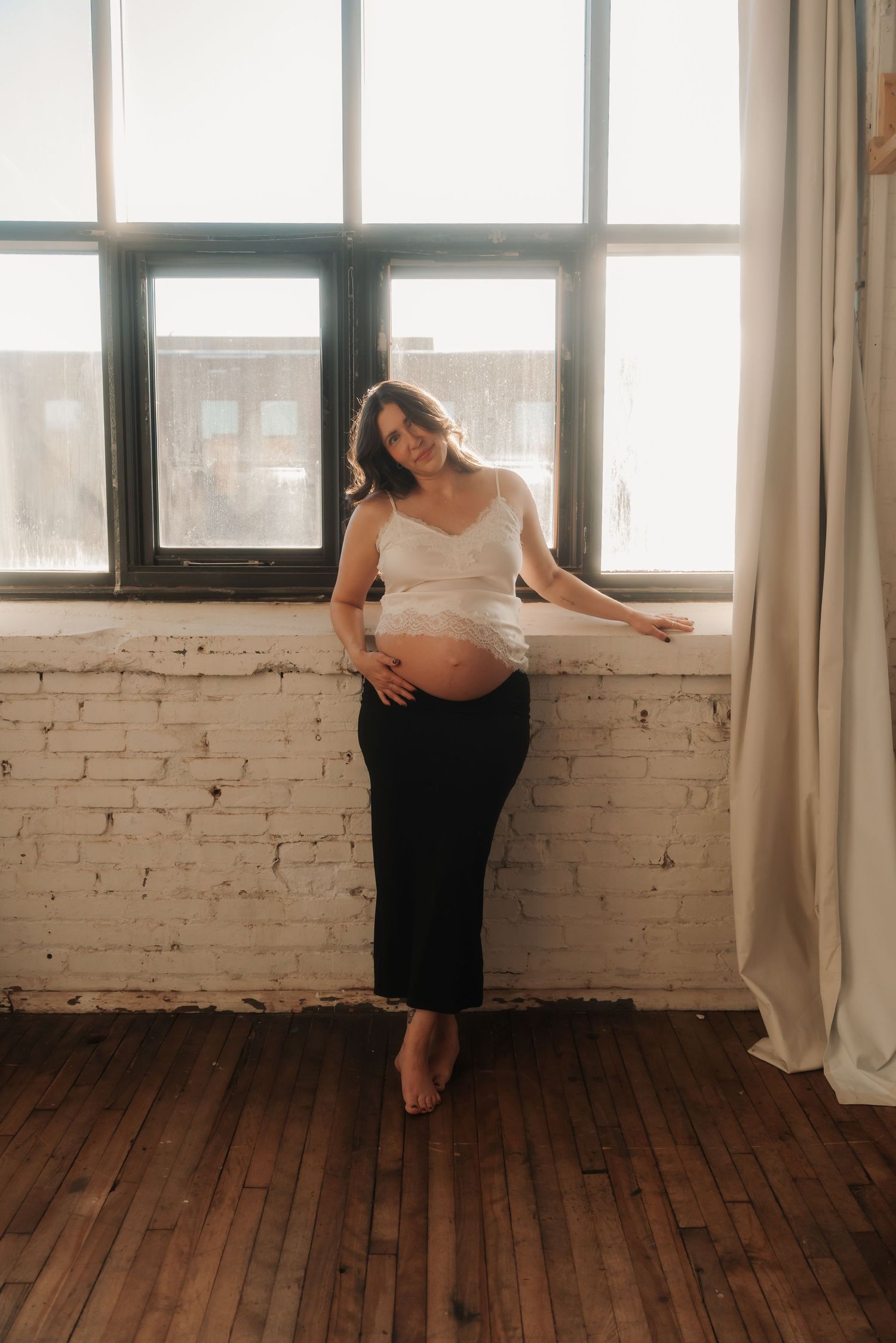 Smiling pregnant woman in white top leaning against window in sunlit loft.