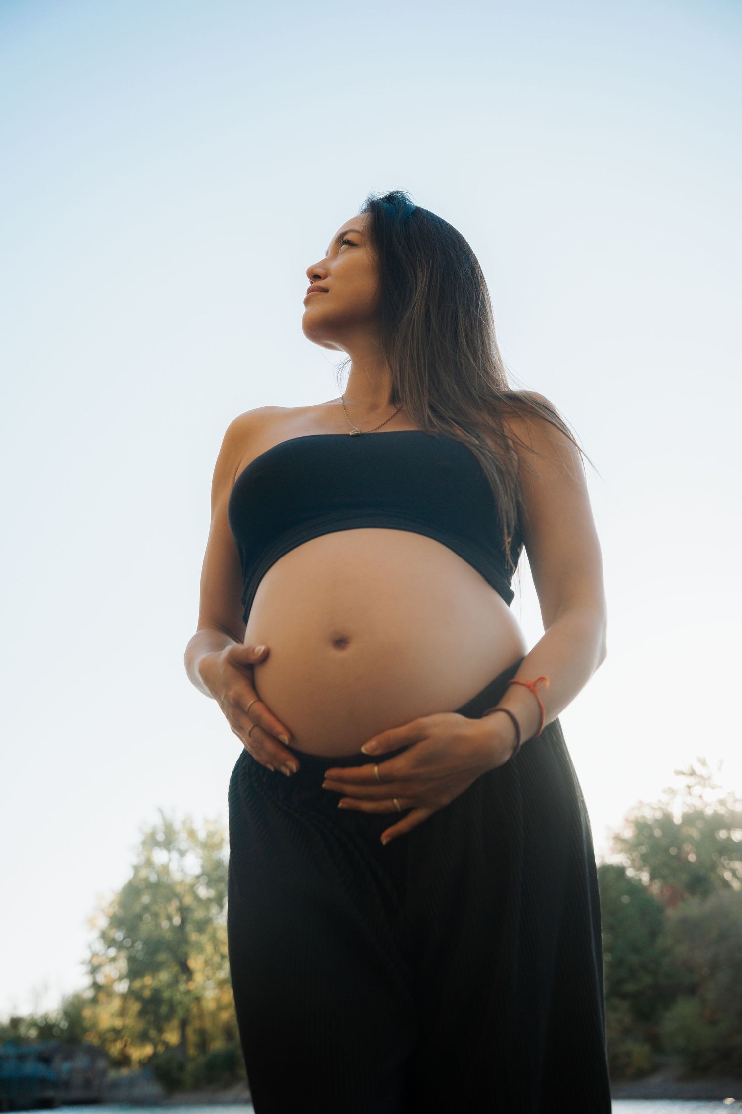 Pregnant woman in black crop top holding her bare belly outdoors against a clear sky in natural sunlight.