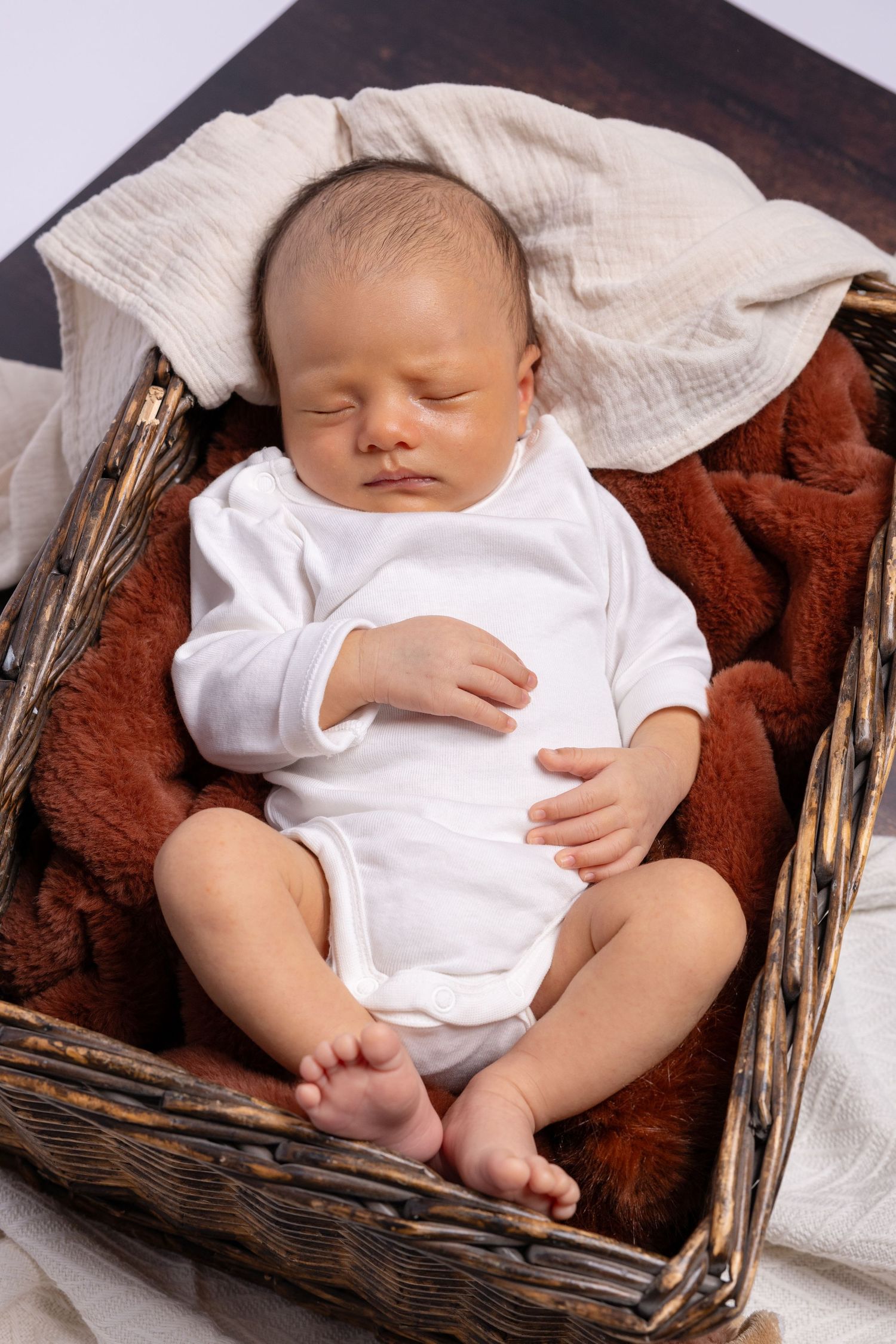 Sleeping baby in white onesie resting peacefully in wooden basket with brown cushioning.