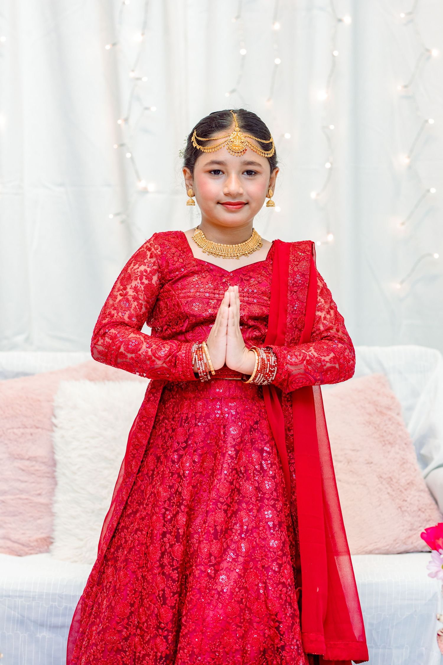 Child in sparkly red dress performing traditional greeting gesture against white backdrop.