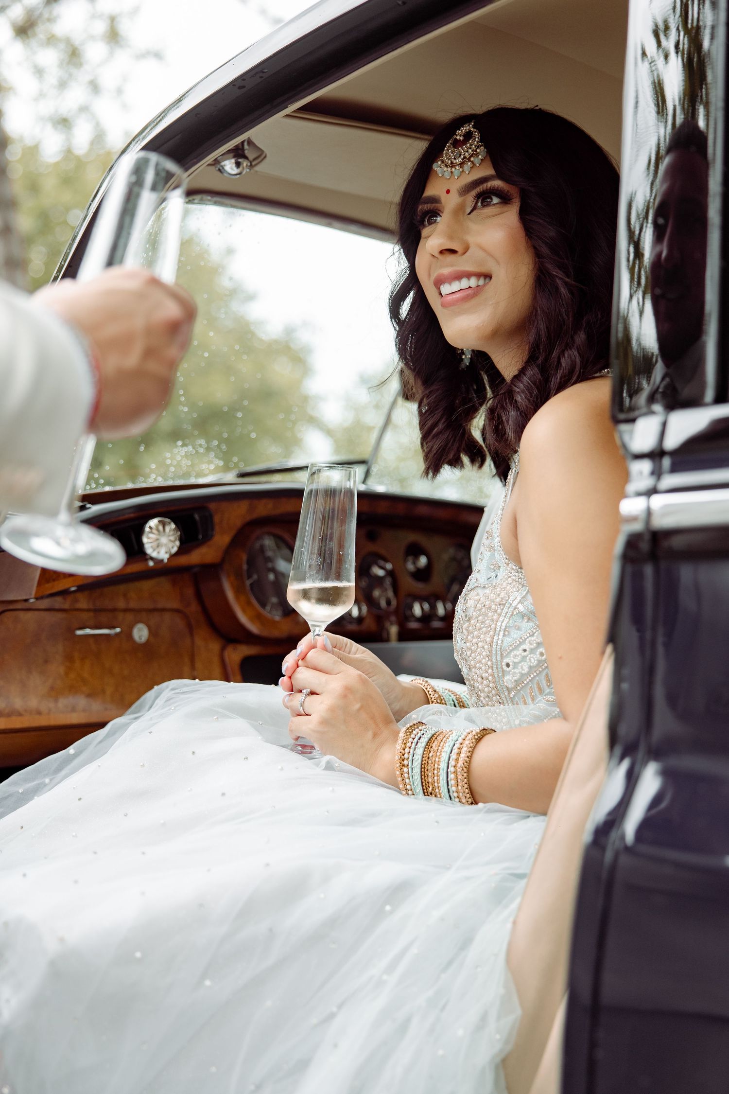 A bride enjoys champagne while seated in a classic car with wooden interior.