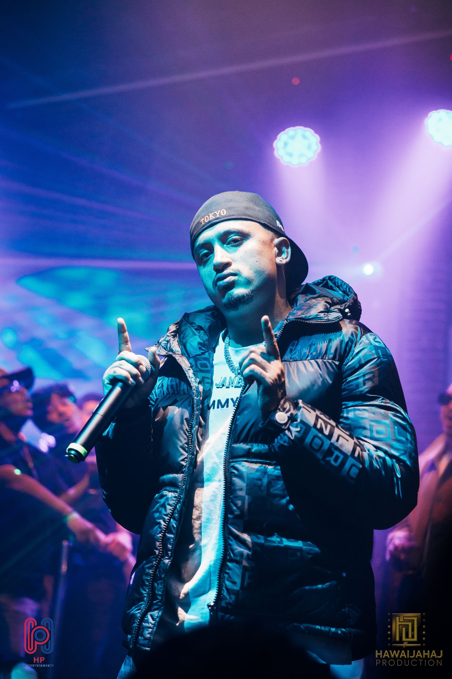 Hip hop artist performing on stage under dramatic purple and blue lighting wearing a puffy black jacket.