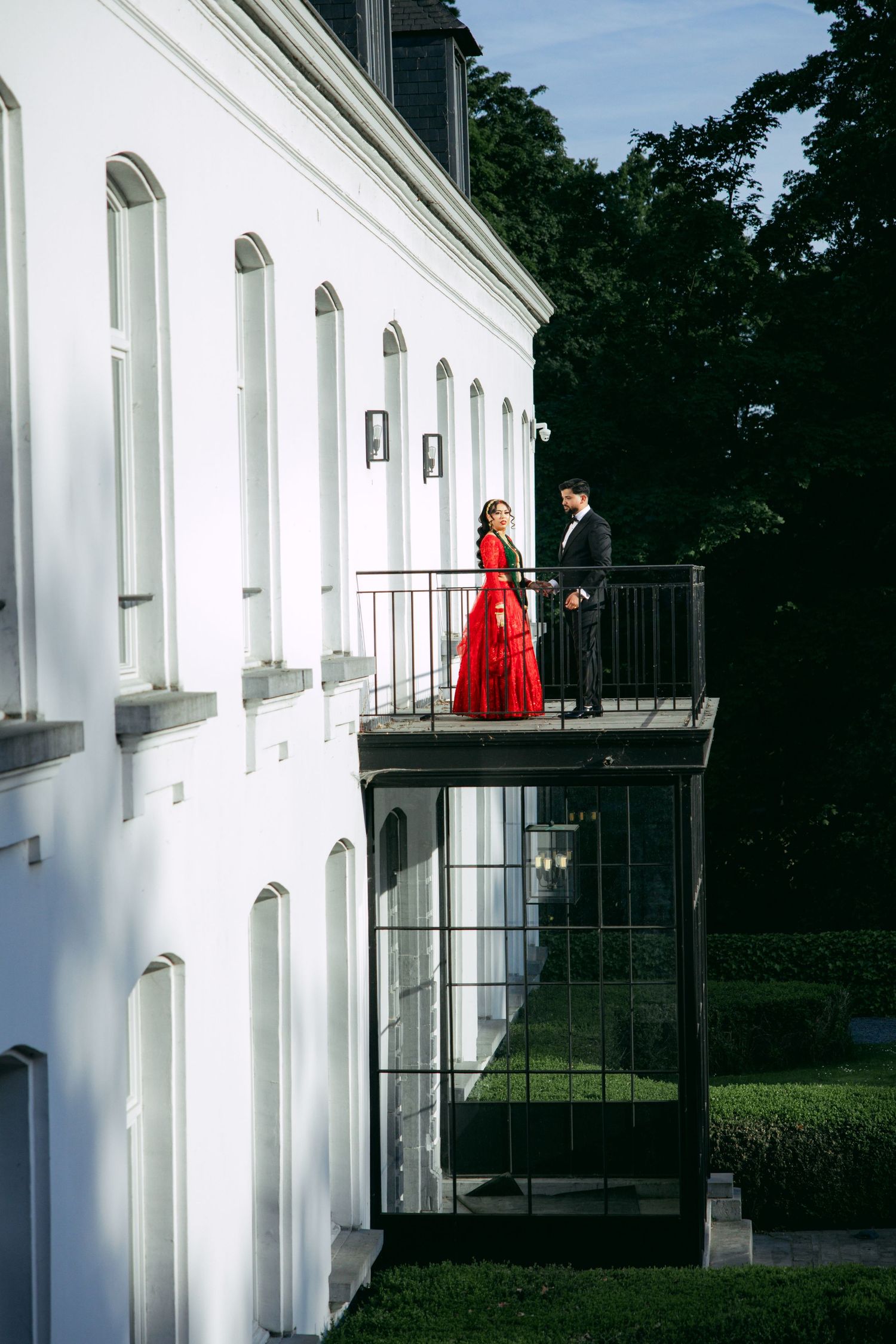 Couple in formal attire stands on white balcony during wedding photoshoot.
