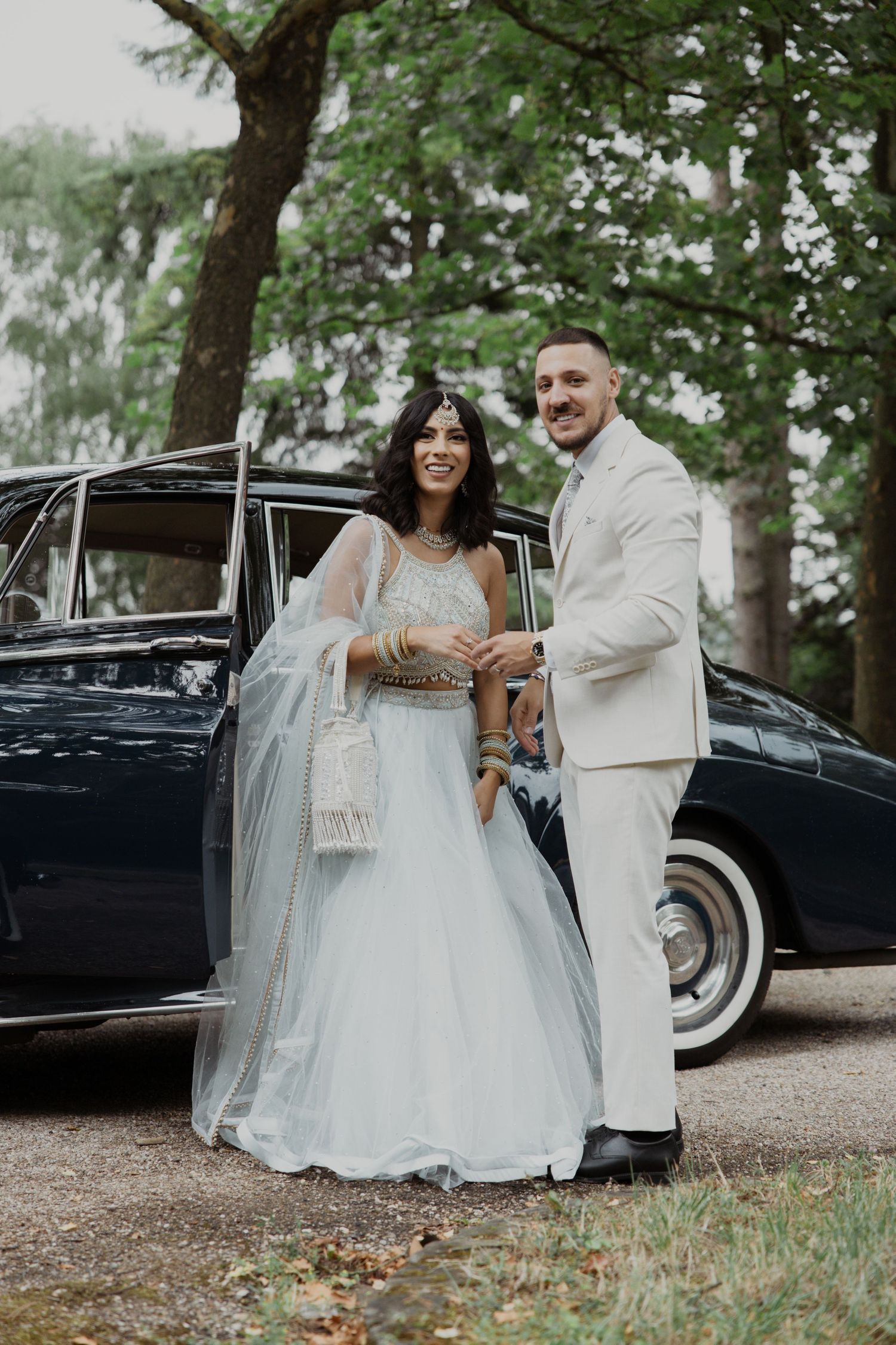 A couple poses near a vintage black car in an outdoor setting with trees.