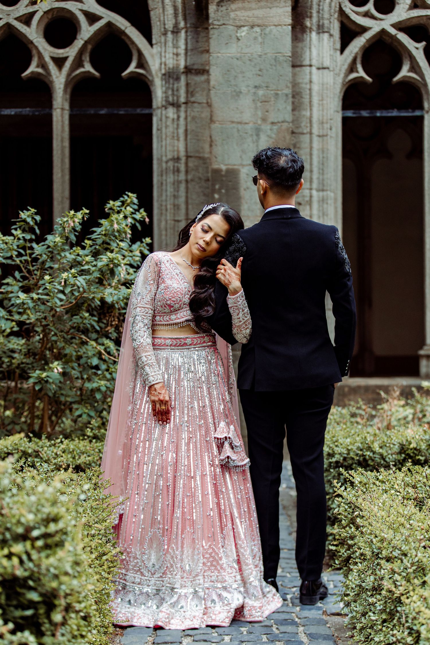 Bride in sparkly pink lehenga poses with groom against gothic architecture backdrop.