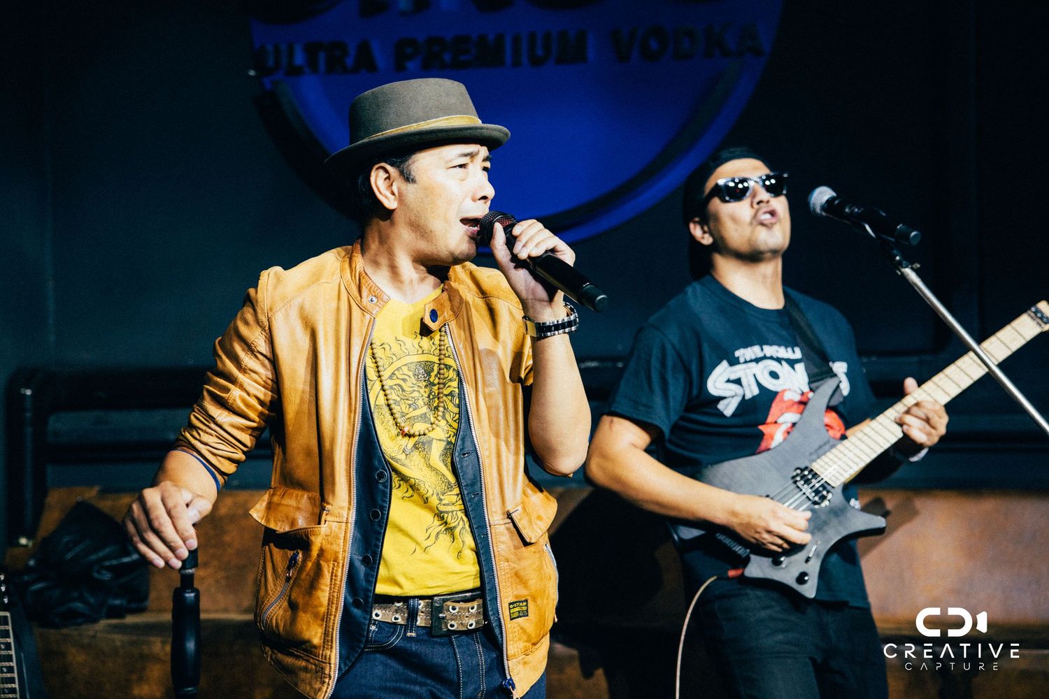 Performers in casual attire singing and playing guitar on stage with blue backdrop.