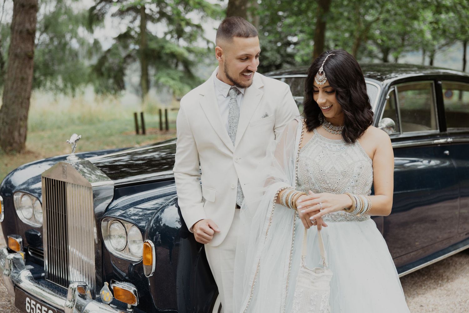 Newlyweds share a moment beside a vintage Rolls Royce in a wooded setting.