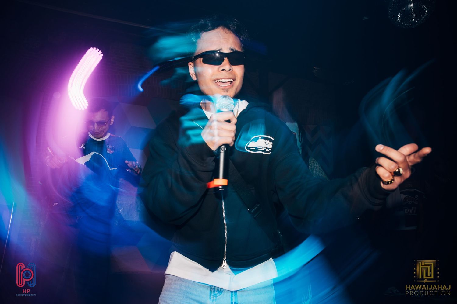 Performer in sunglasses and jacket singing on stage with colorful purple lighting effects in nightclub setting.