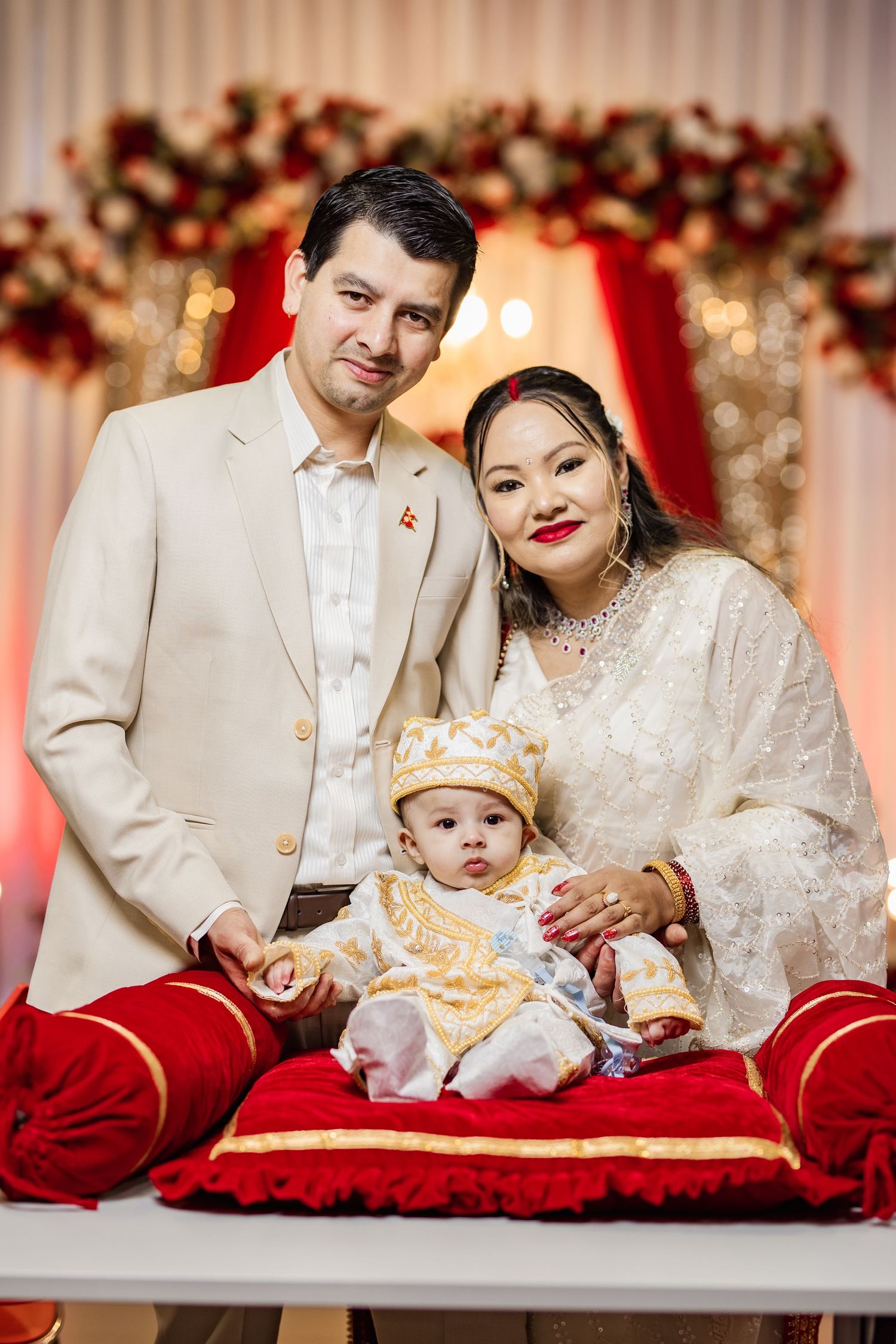 Family portrait with baby in festive attire against red and white decorated backdrop.
