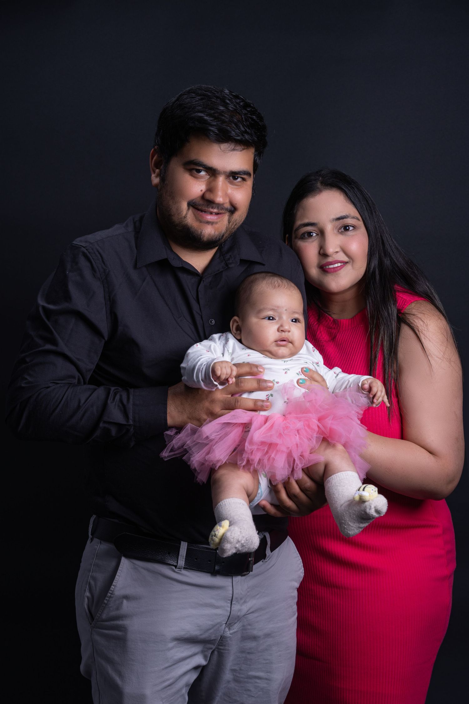 Family portrait against dark background with baby in pink tutu dress.