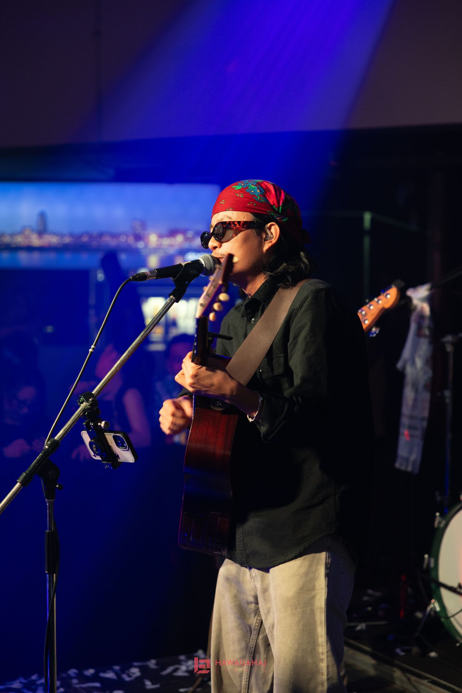 Performer playing acoustic guitar on stage with blue spotlight and cityscape background.