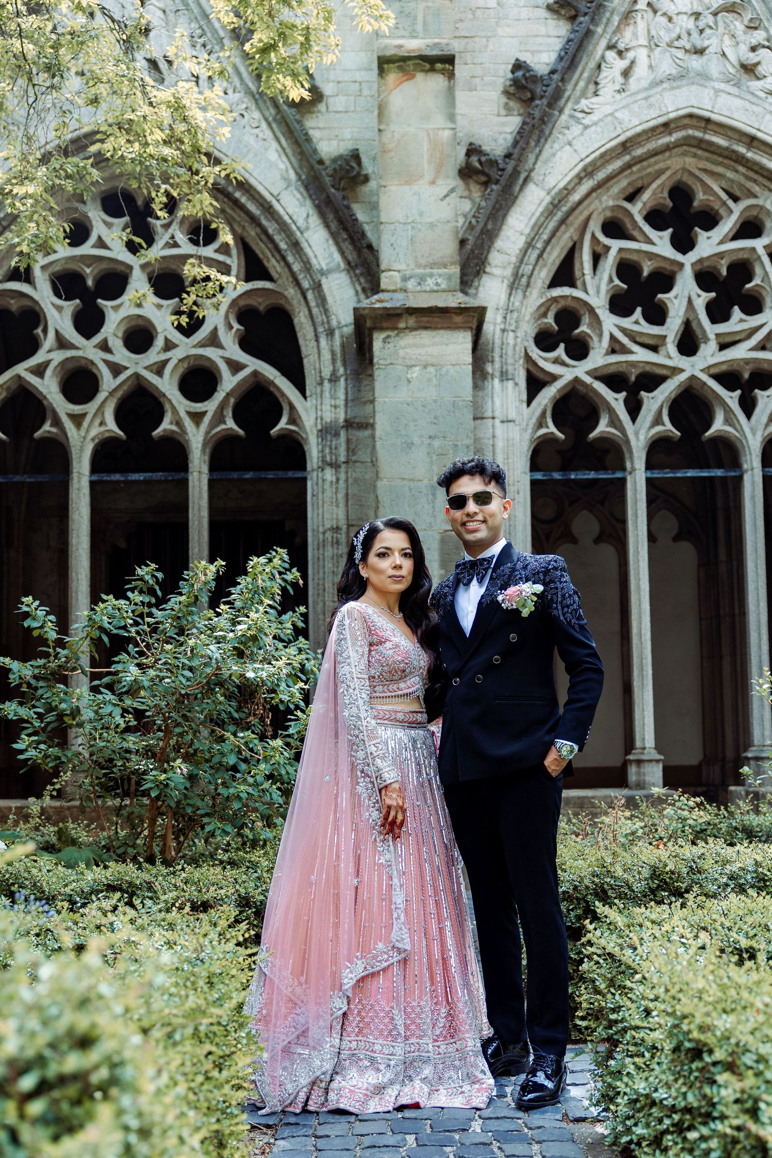 A couple poses in formal attire against ornate gothic architecture and garden backdrop.