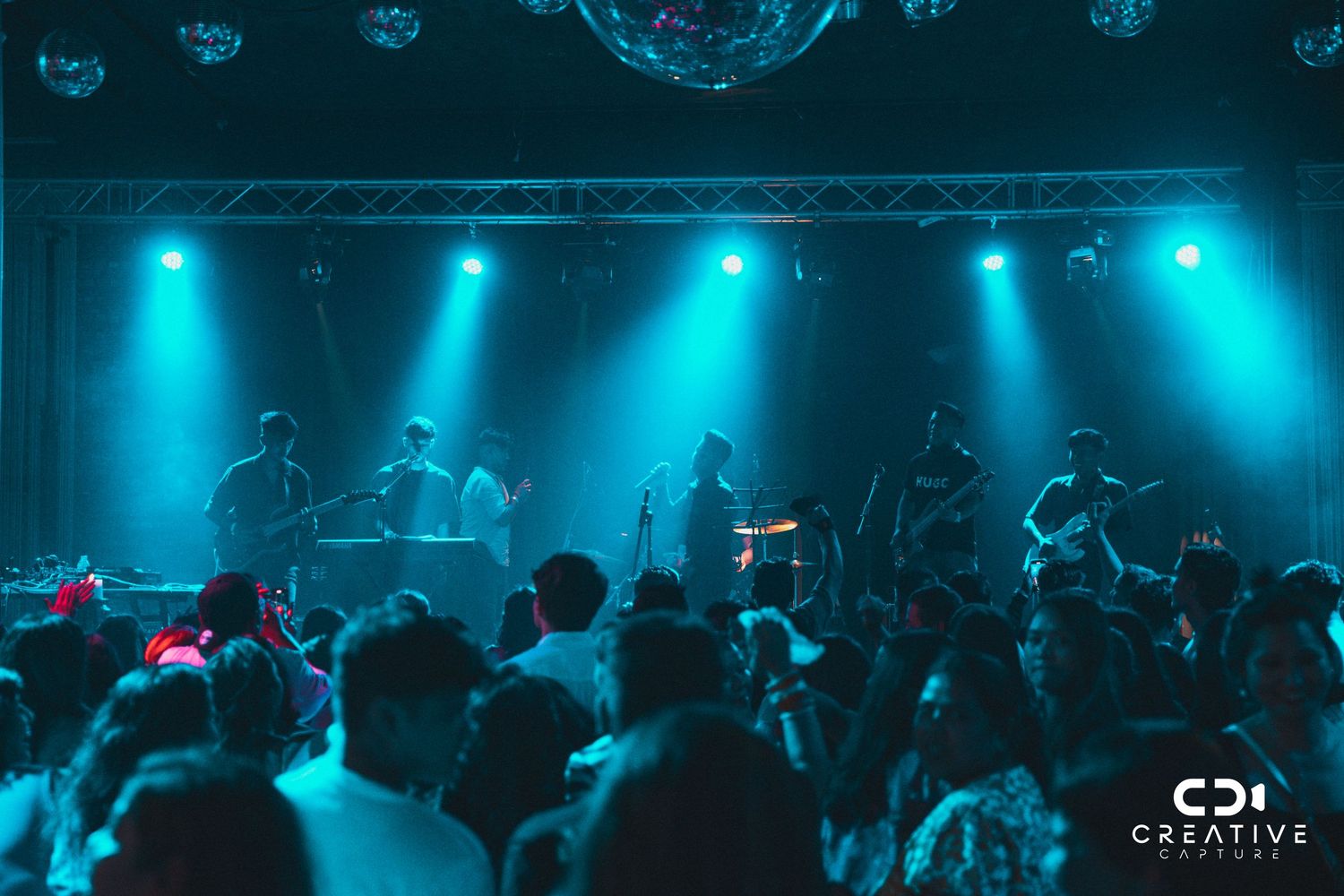Concert venue with teal lighting and silhouetted crowd watching performers on stage.