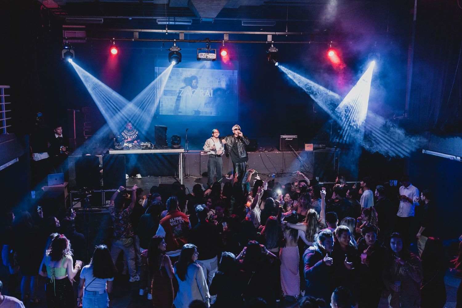 Crowded nightclub venue with dramatic crossing spotlights illuminating the dance floor.