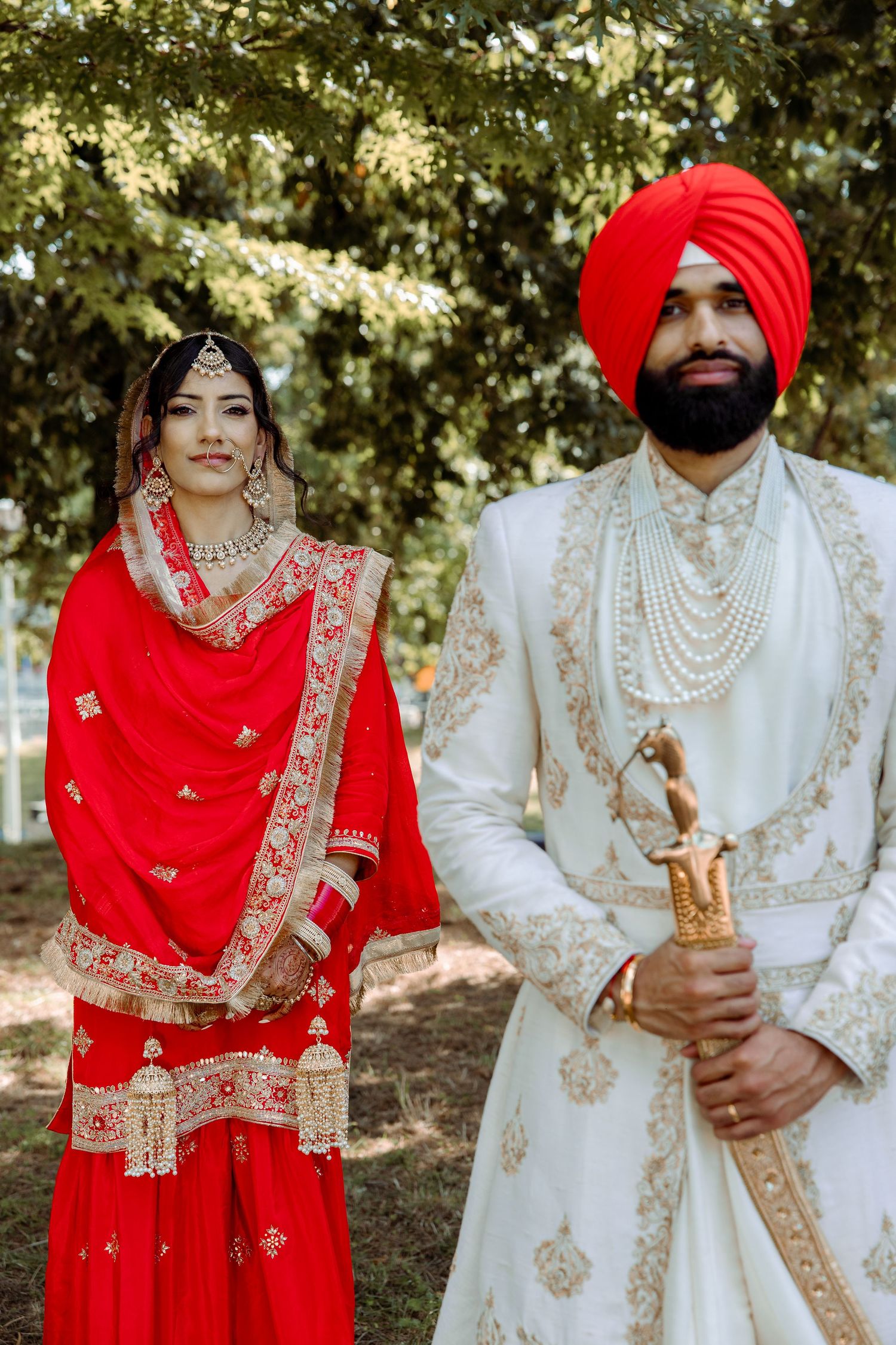 Traditional Sikh wedding couple in ornate red and white outfits pose together outdoors.