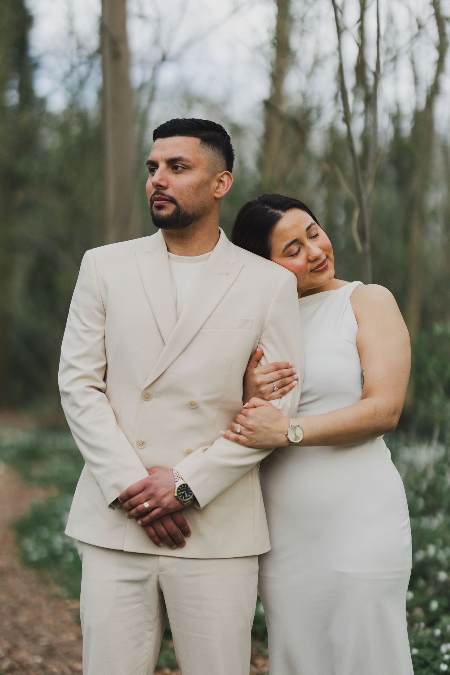 A couple in cream-colored outfits shares an intimate moment in a wooded setting.