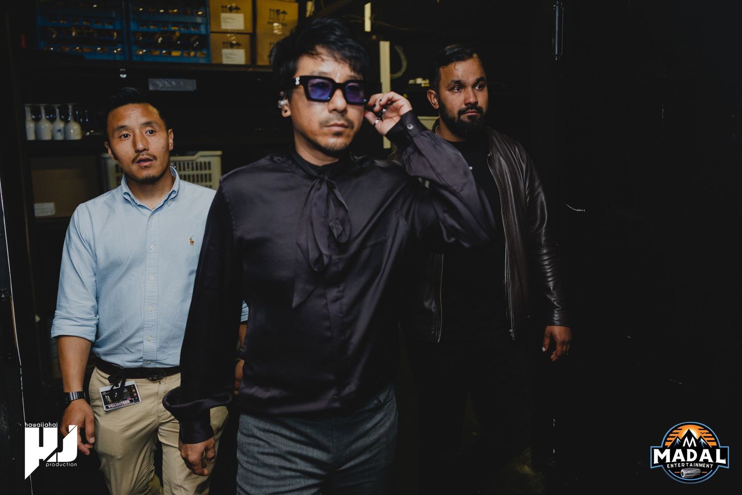 Three performers walking backstage in dark attire with one wearing sunglasses.