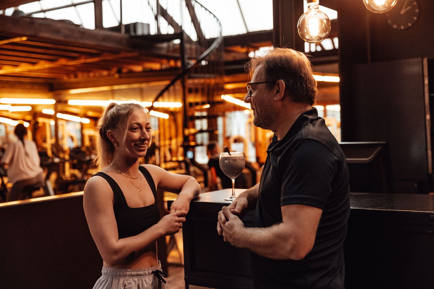 People socializing in a warmly lit industrial-style fitness facility.
