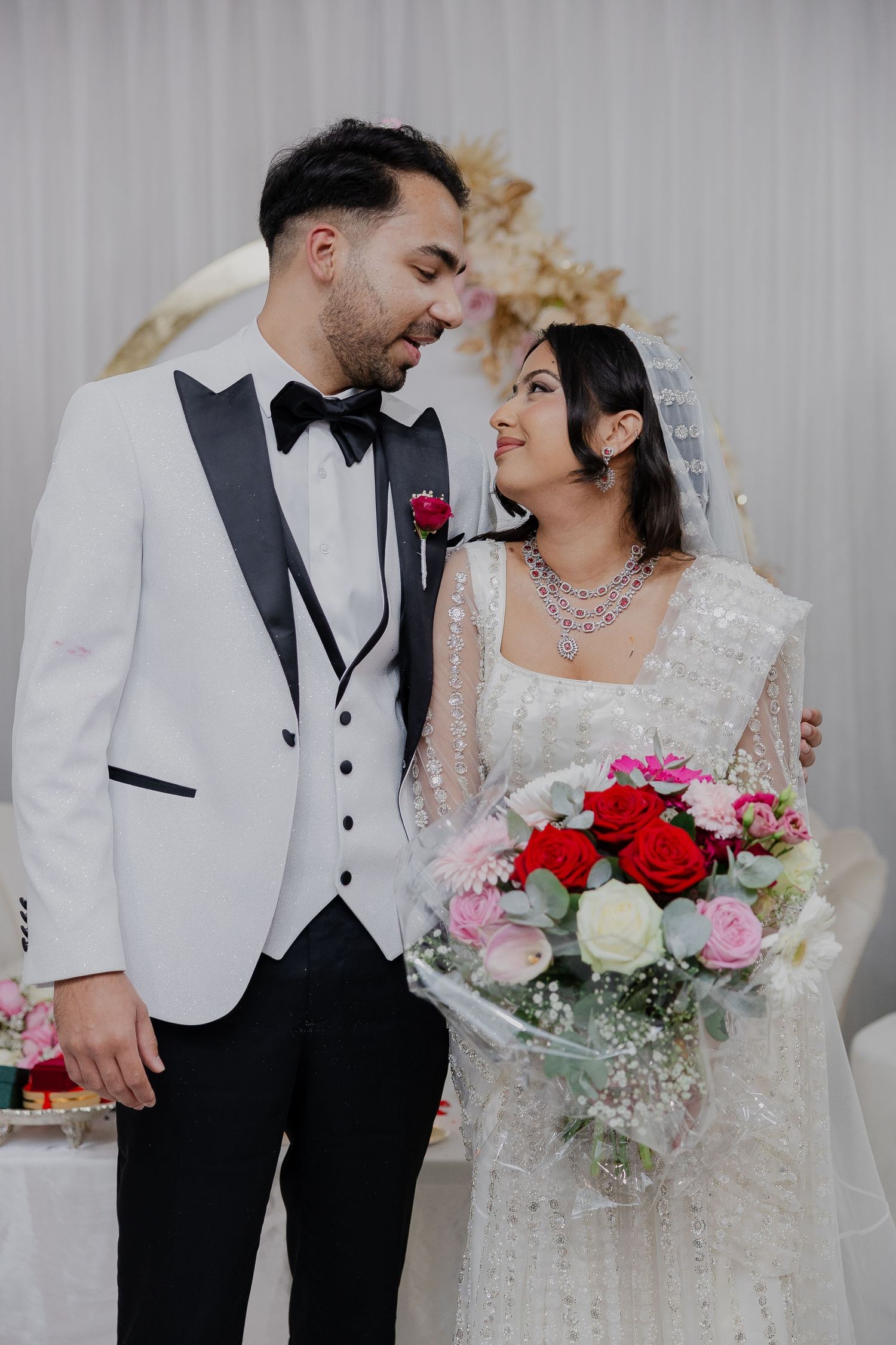 A romantic wedding portrait featuring a white tuxedo and lace dress with red and pink roses in the bouquet.