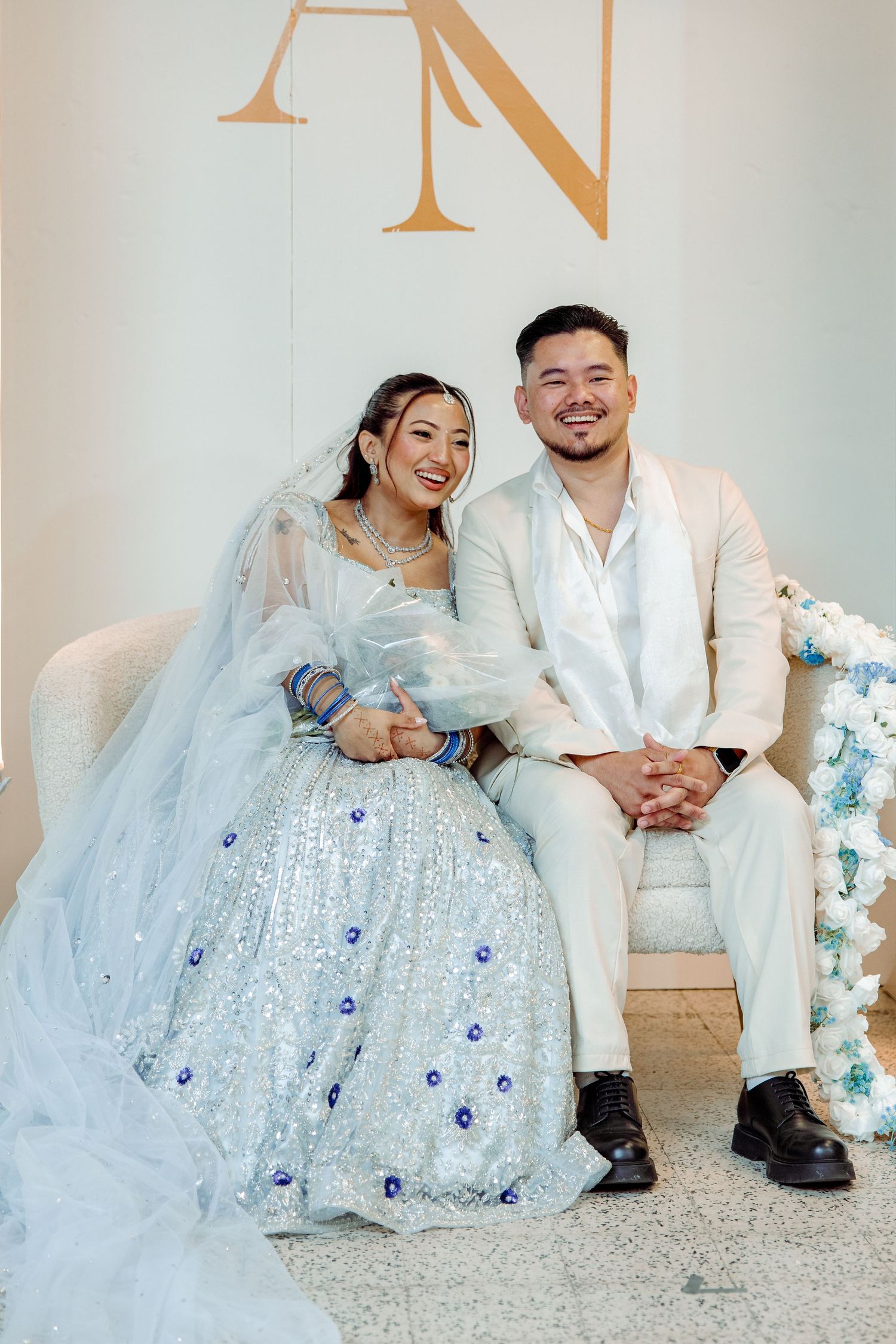 Newlyweds in white attire sit together on couch under monogram wall decor.