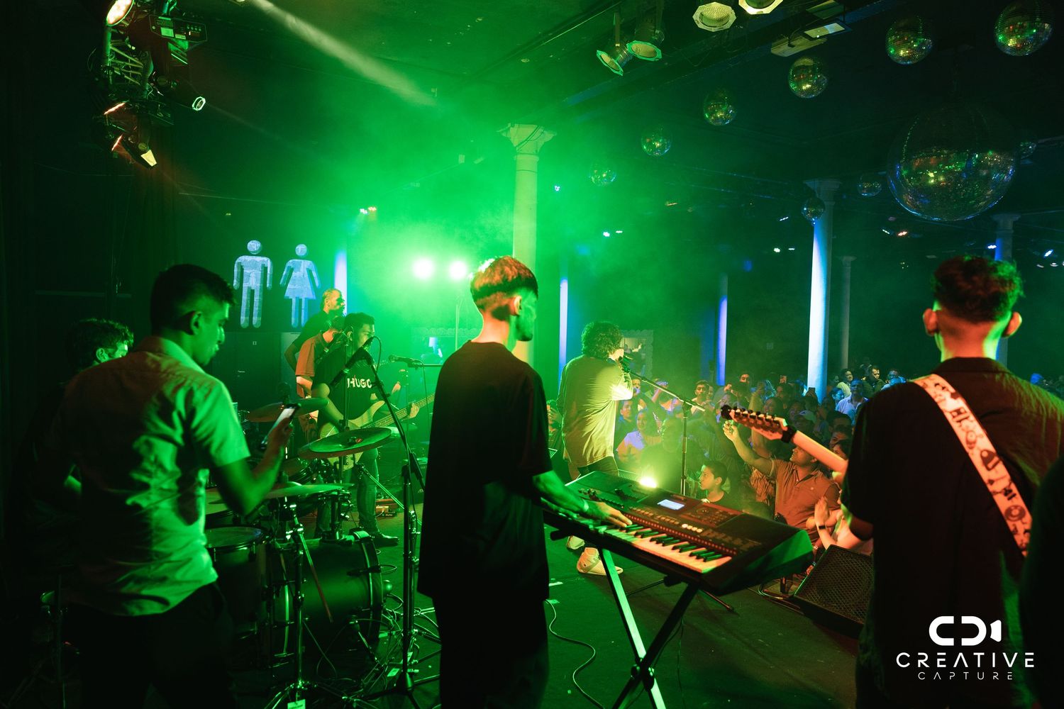 Band performing on stage with green lighting and electronic equipment setup.