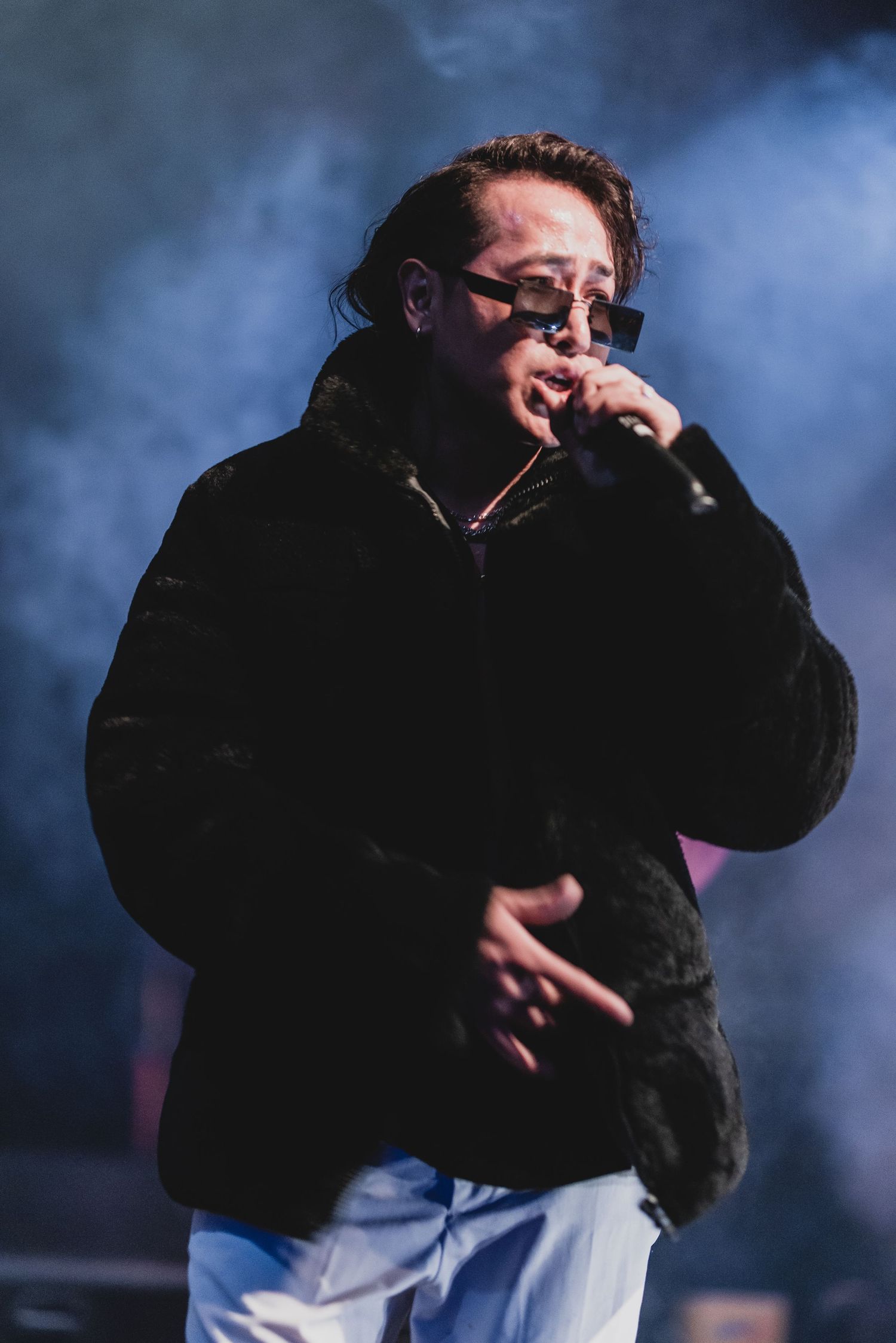 Performer in black coat and sunglasses on stage with dramatic blue smoke effects in the background.
