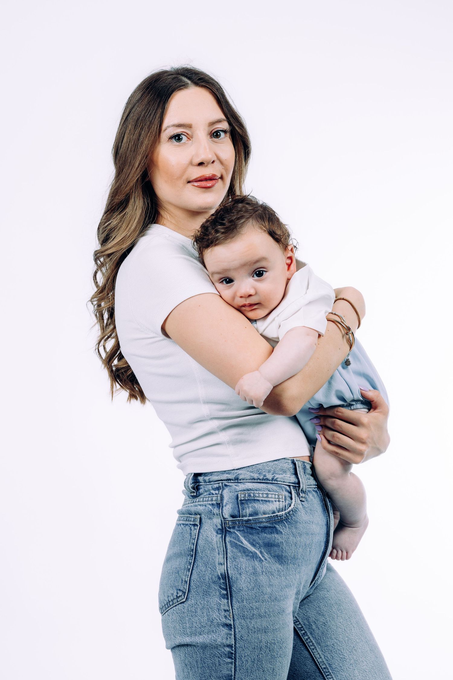 Mother in casual wear holds and embraces baby against white studio backdrop.