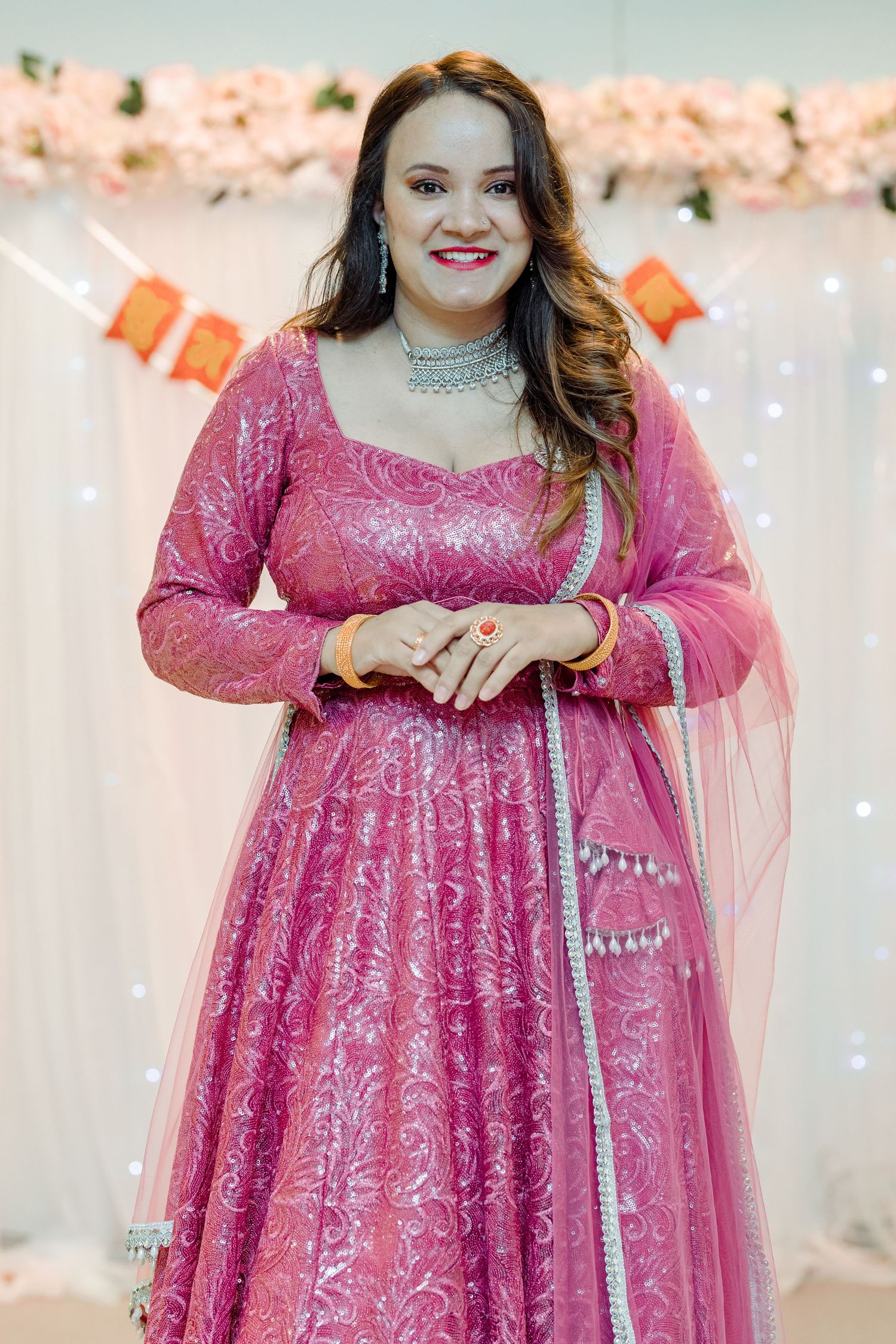Woman in sparkly pink formal gown posing against white backdrop with string lights.