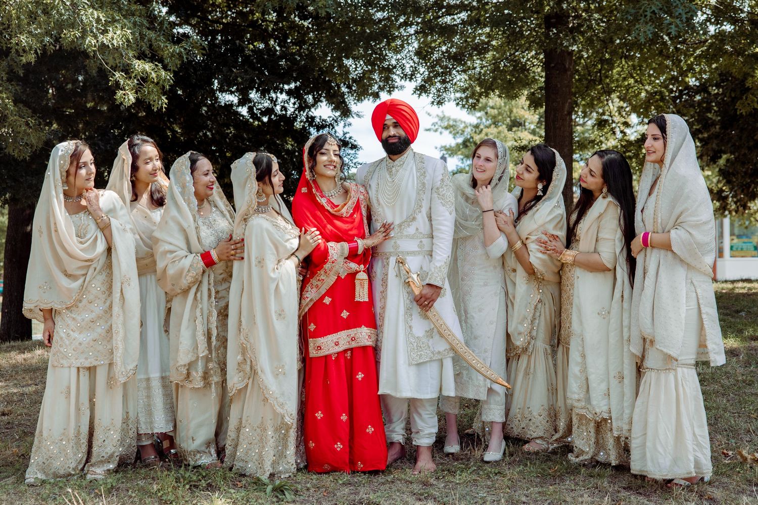 Large wedding party in traditional Indian attire poses outdoors in white and red colors.
