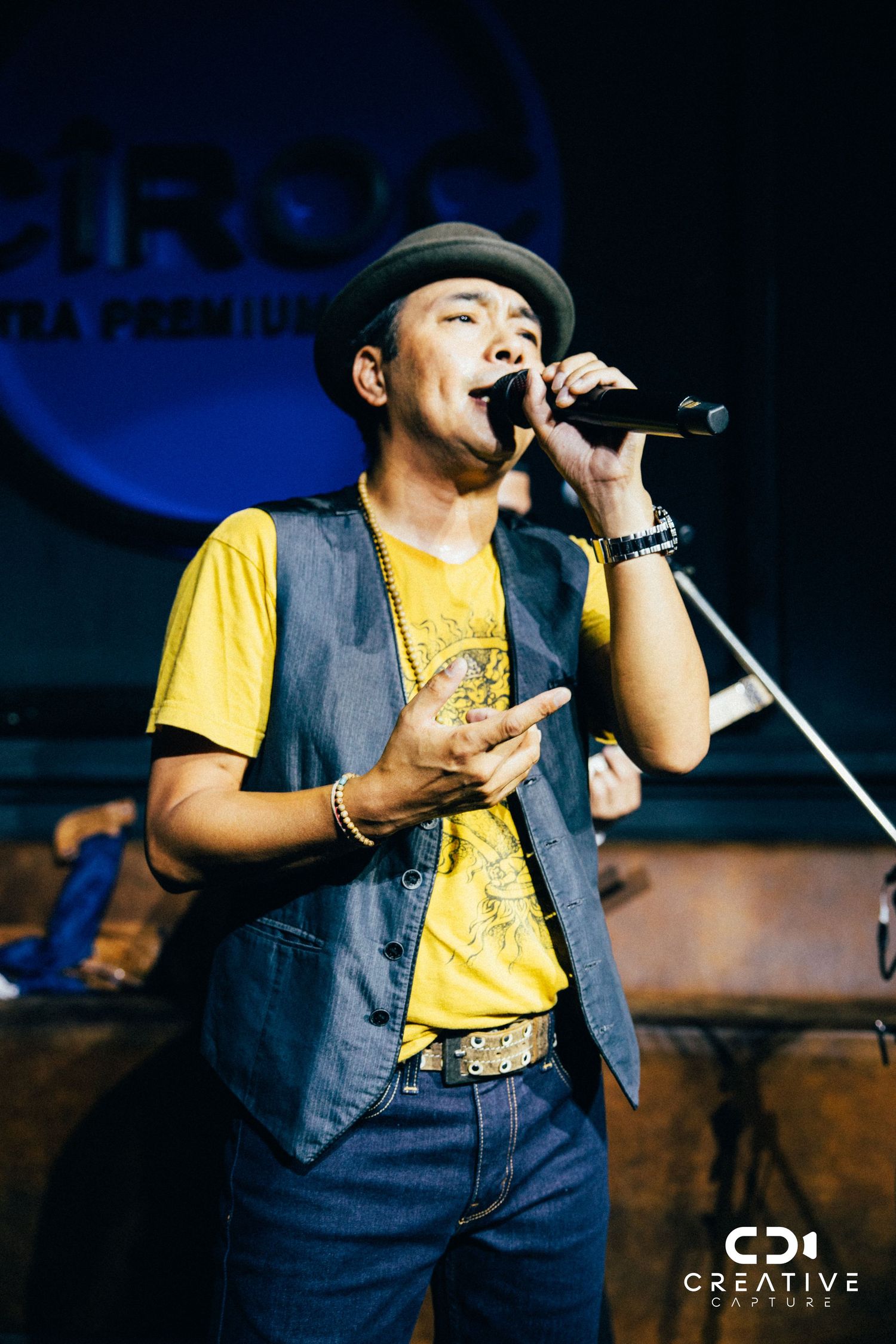 Performer in yellow shirt and denim vest singing passionately on stage.