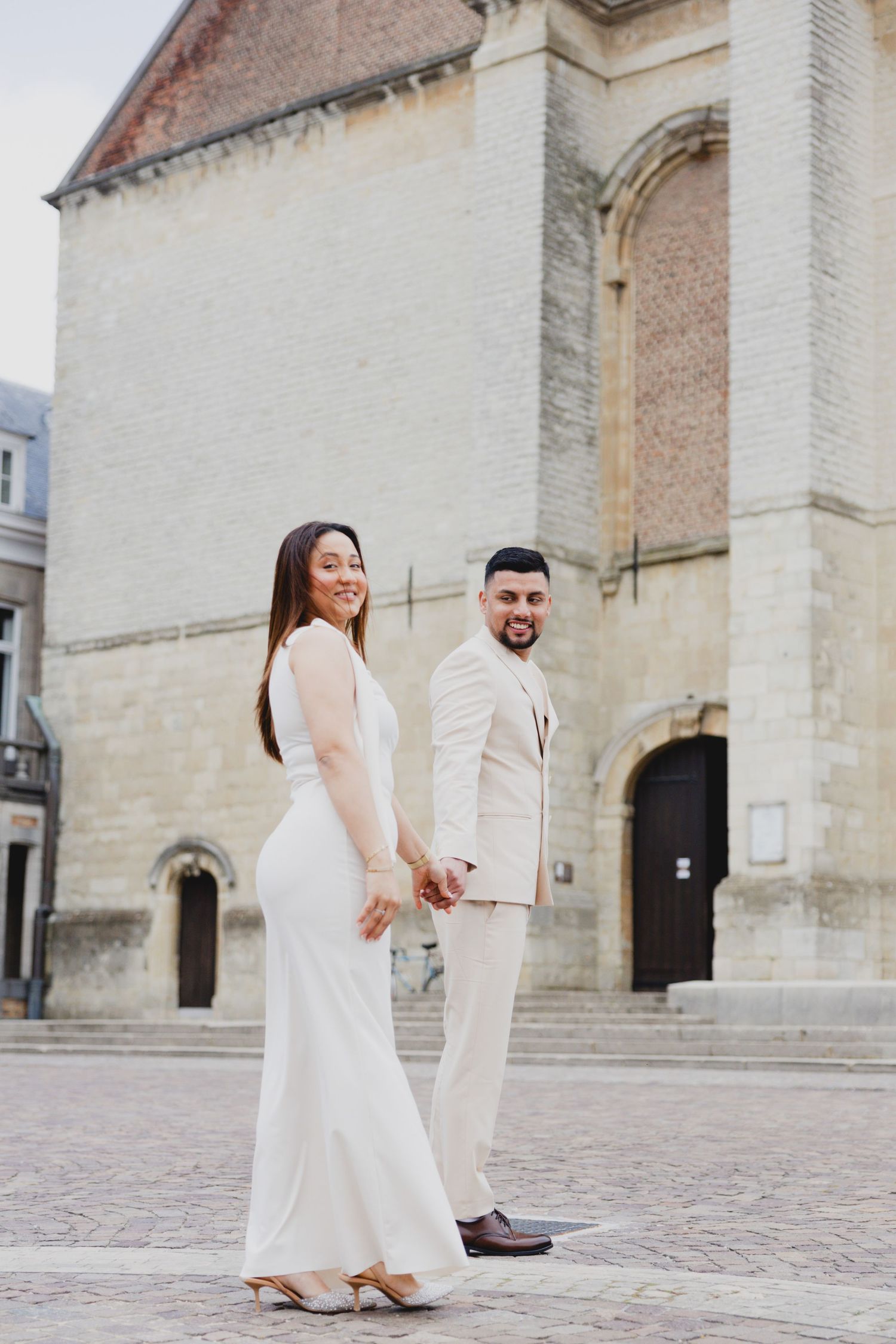 Elegant couple in white attire posing for wedding photos in front of a historic stone church.