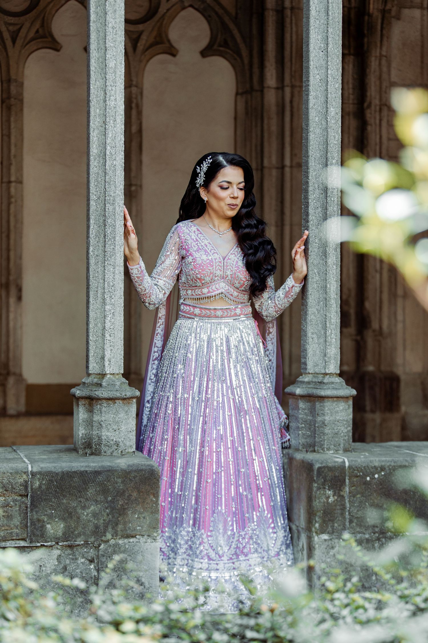 A bride in a sparkly pink lehenga poses elegantly between gothic stone pillars.
