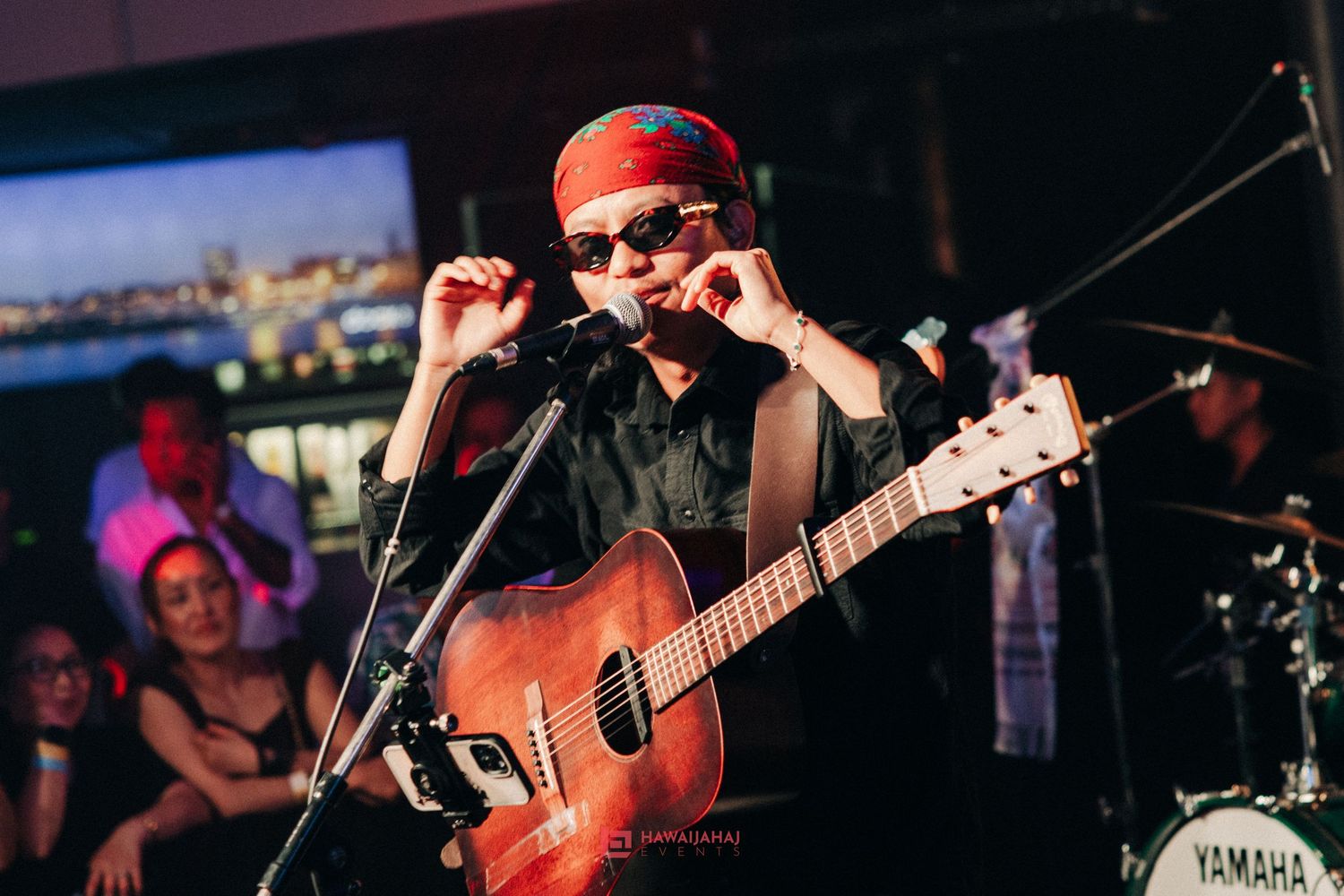 Musician performing with acoustic guitar on stage under spotlight with city backdrop.