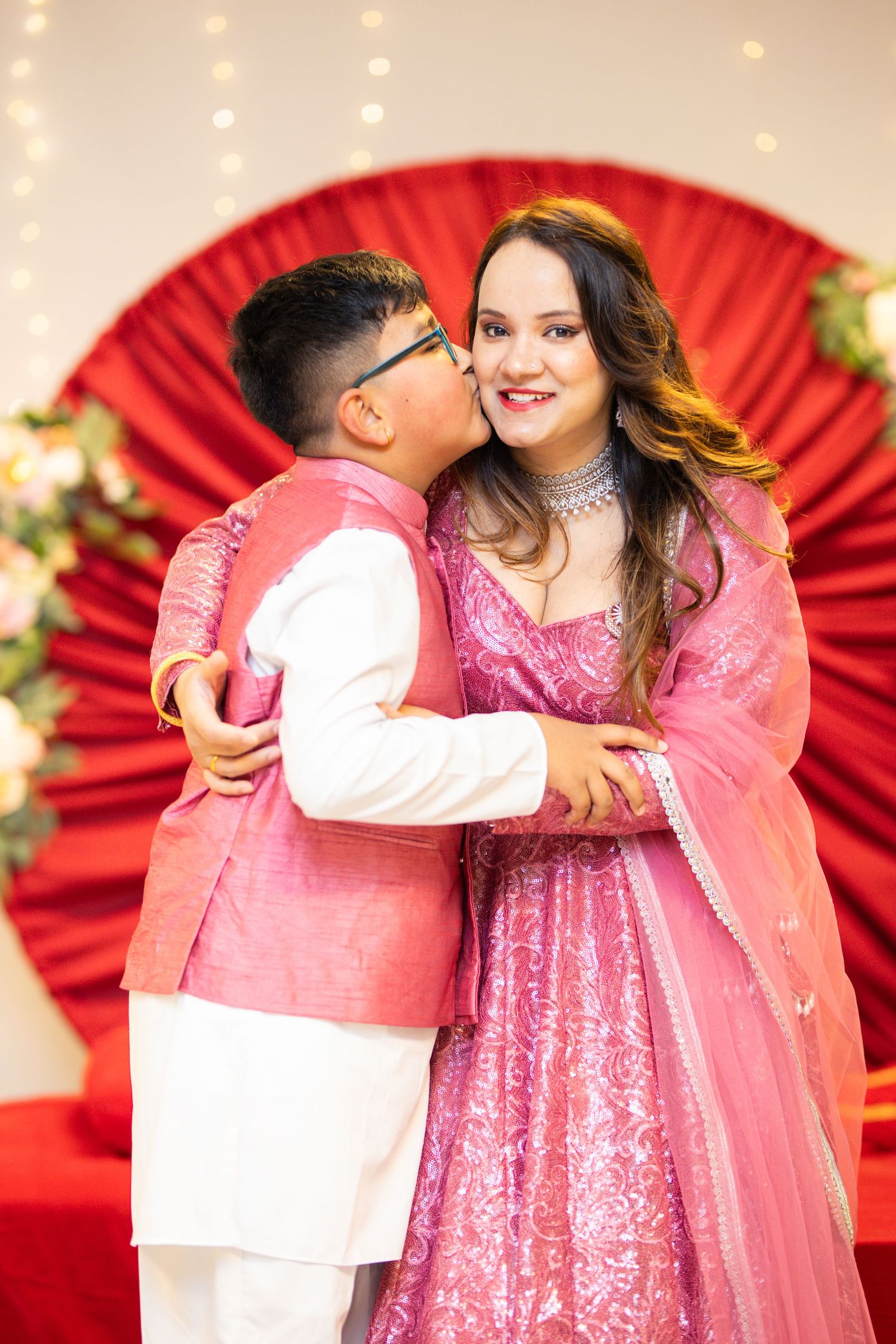 Romantic couple in pink and white outfits embracing against red backdrop with string lights.