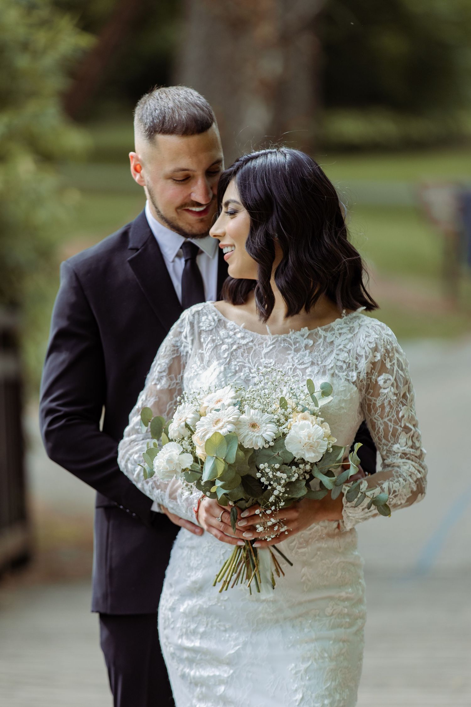 Wedding couple with white floral bouquet poses in formal garden setting.