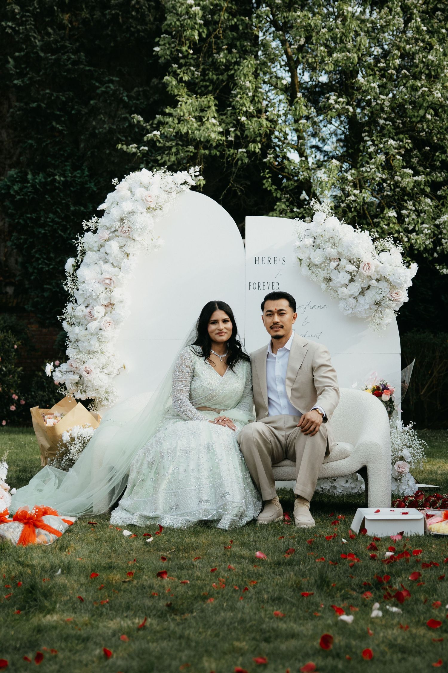 A romantic wedding photoshoot setup with white floral decorations and red rose petals scattered on grass.