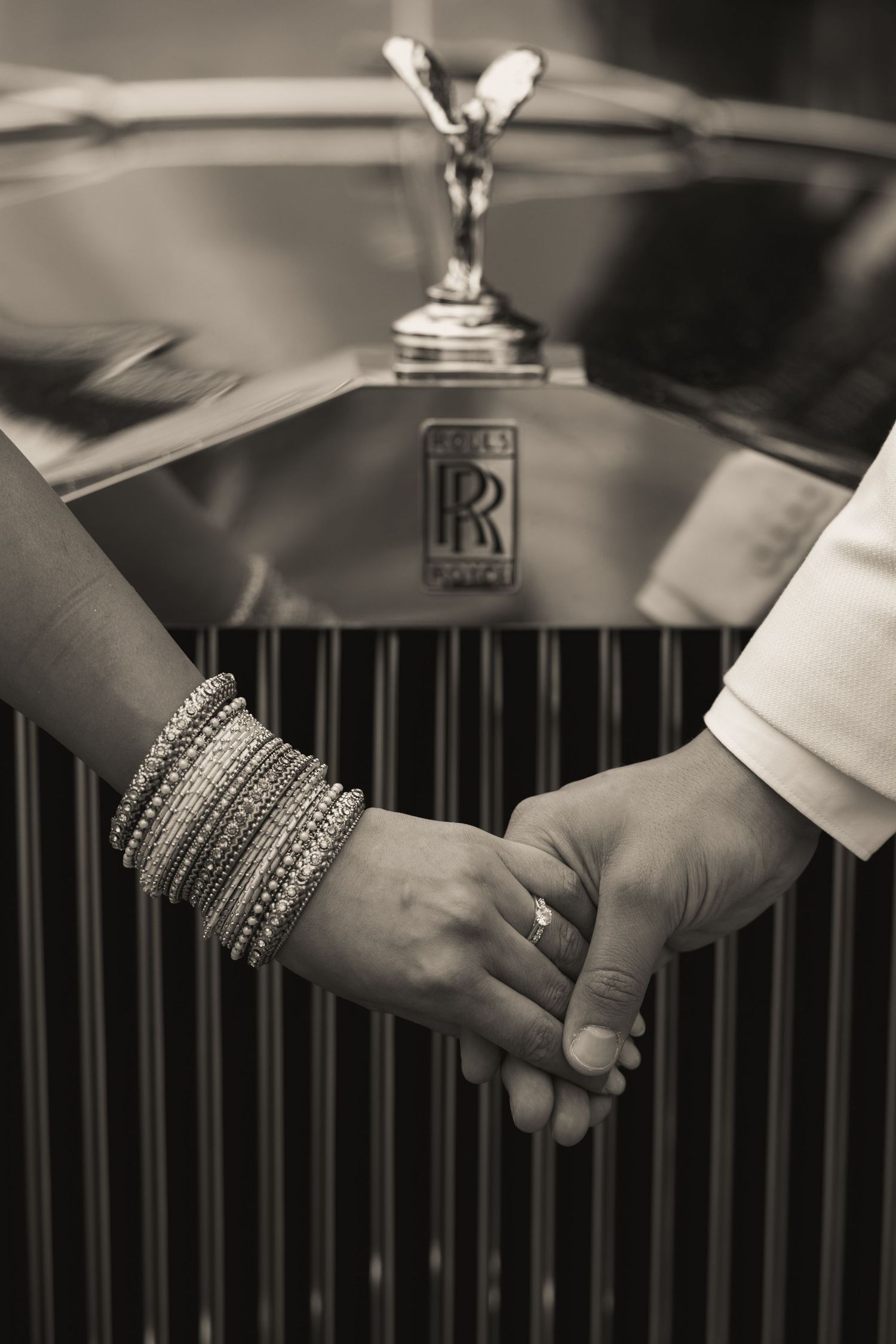 Close-up of hands with jewelry against a Rolls Royce grille in black and white.