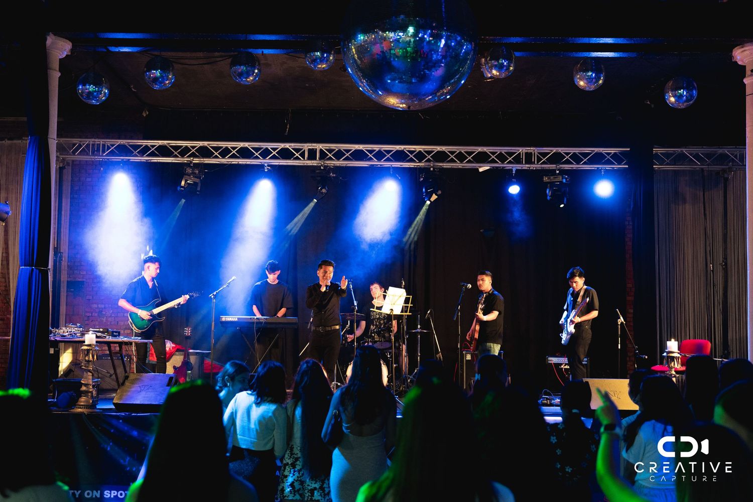 Band performing on stage with blue lighting and crowd silhouettes in foreground.