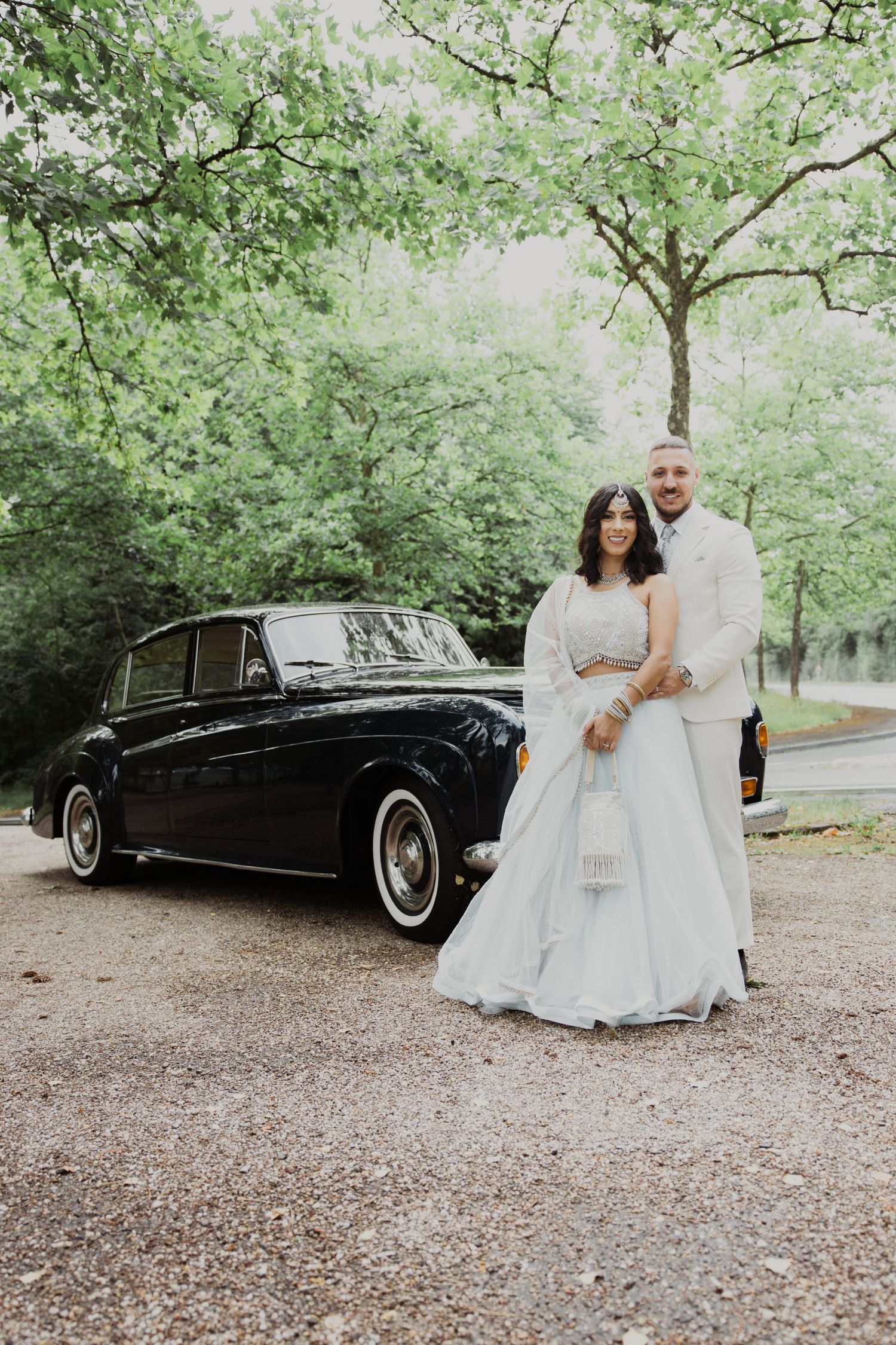 Wedding couple poses in front of vintage black car on tree-lined path.